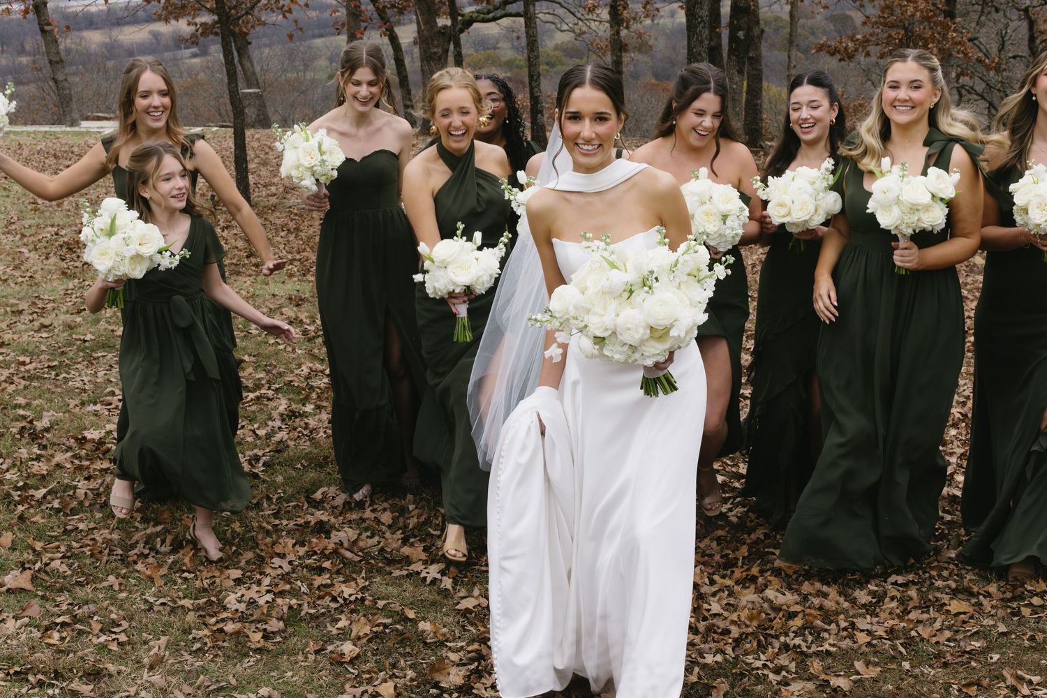 Wedding party in black dresses watches bride in white gown during outdoor autumn ceremony.