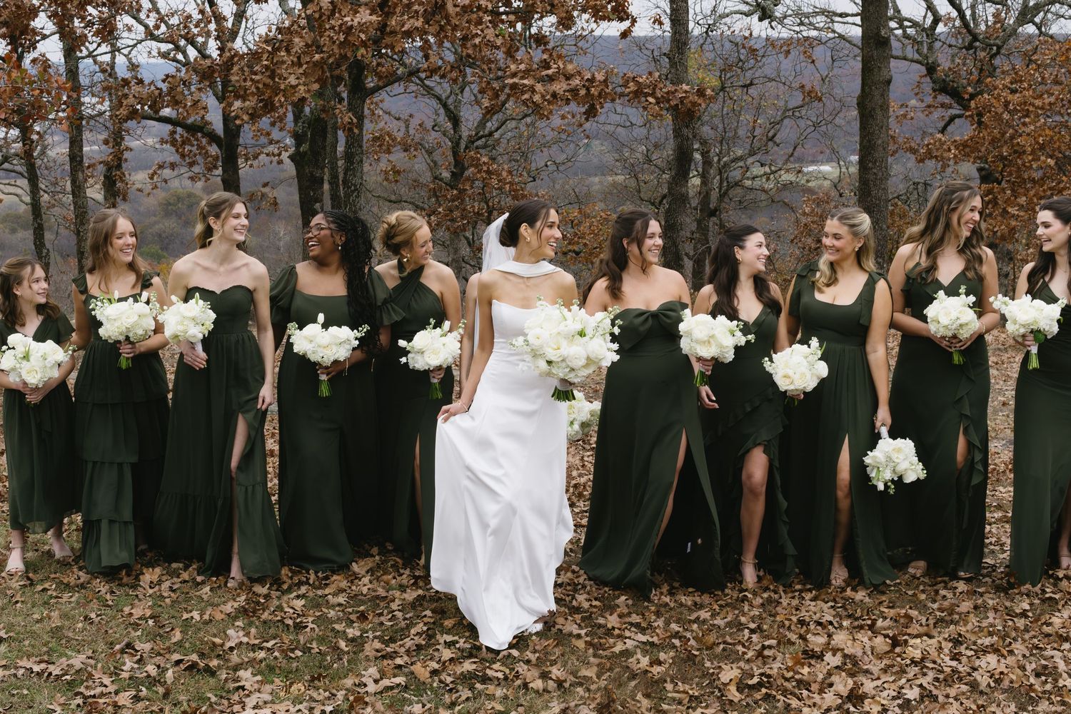 Bridal party poses in line against autumn trees with contrast of black dresses and white bouquets.