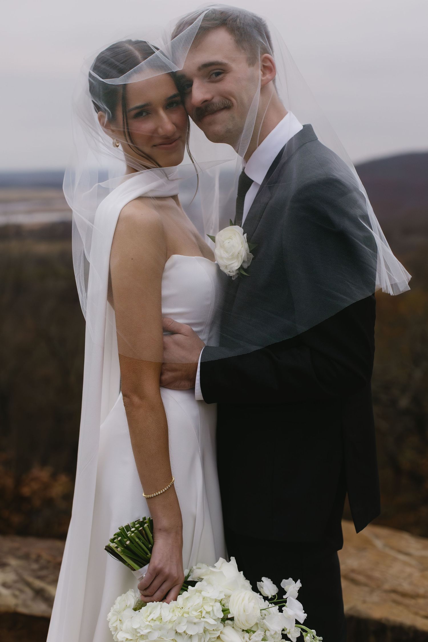 A romantic outdoor wedding photo showing a couple embracing against an autumn landscape backdrop.