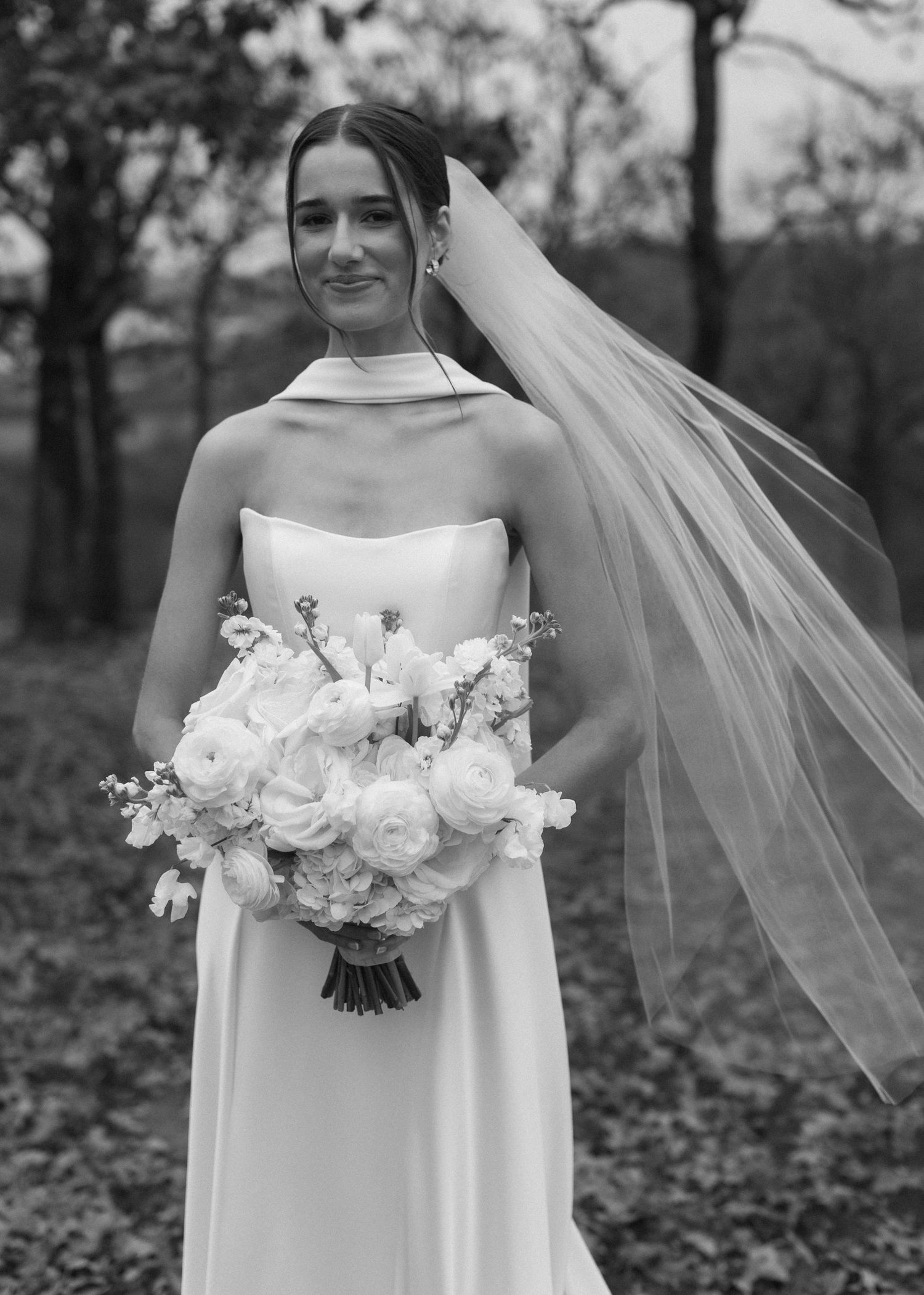 A bride in a strapless white gown holds a white rose bouquet against an autumn backdrop.