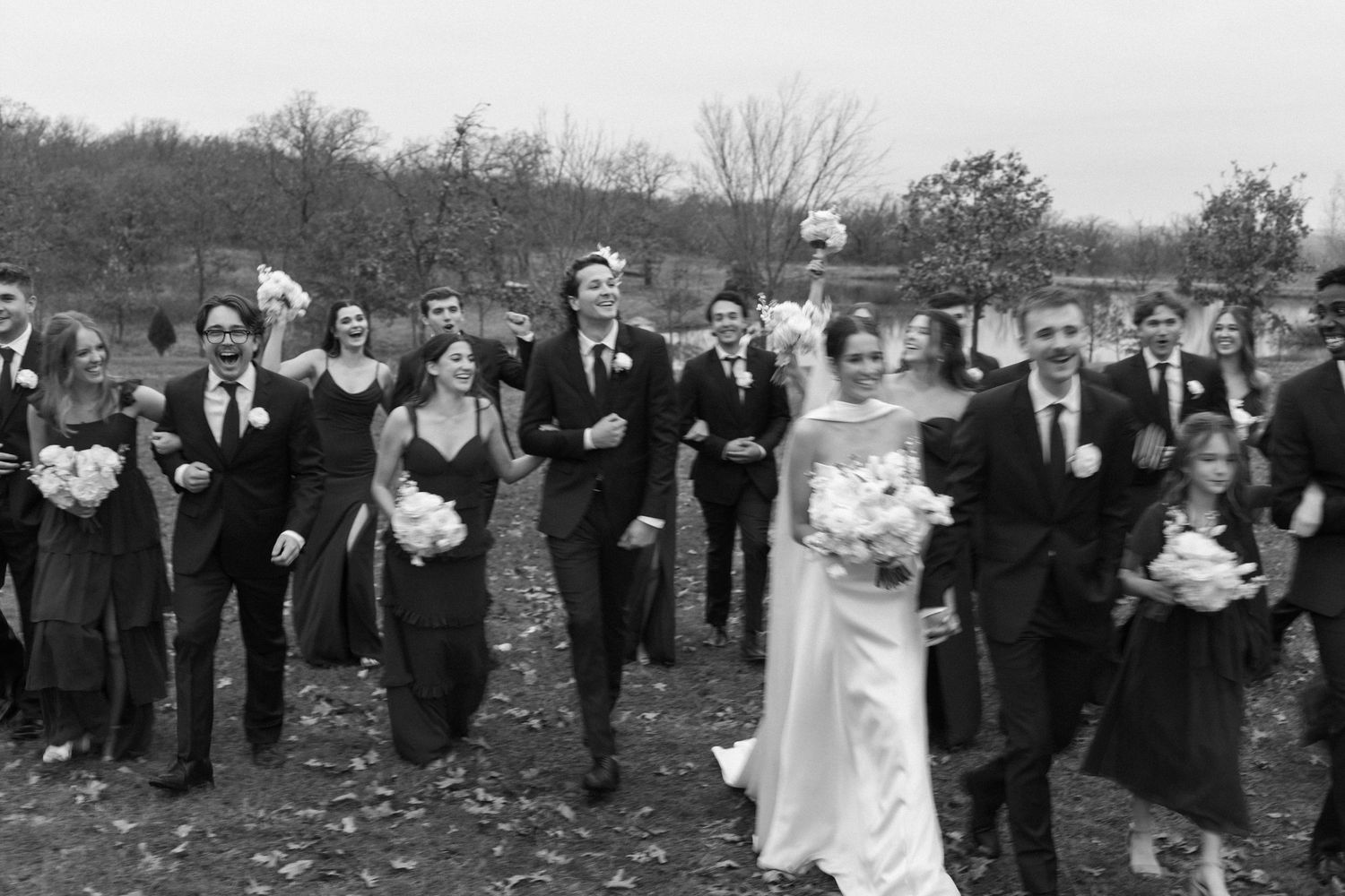 Wedding guests in formal attire gather for a large group photo outdoors.