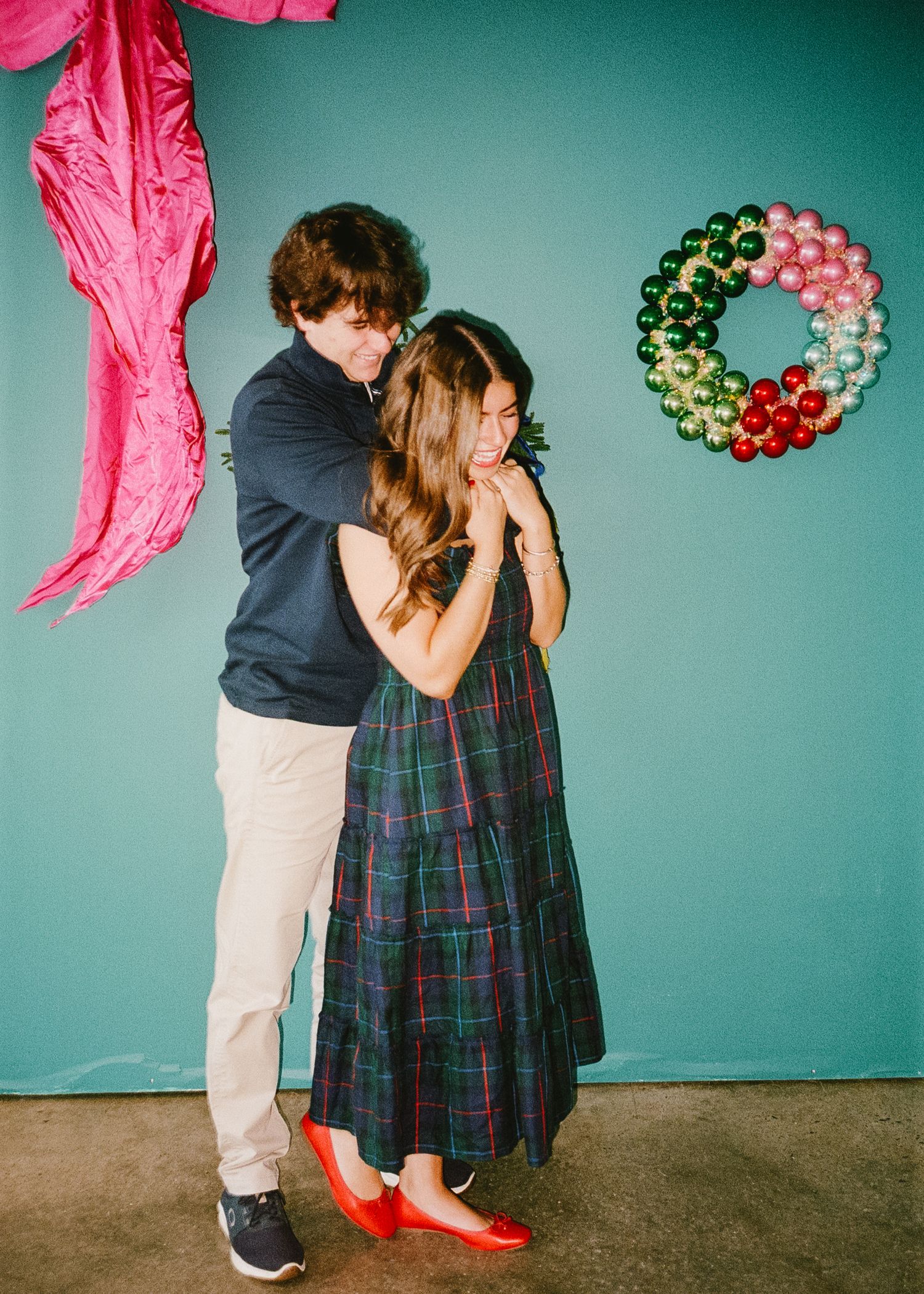 A couple embraces in front of a teal wall with pink decorations and festive wreaths.
