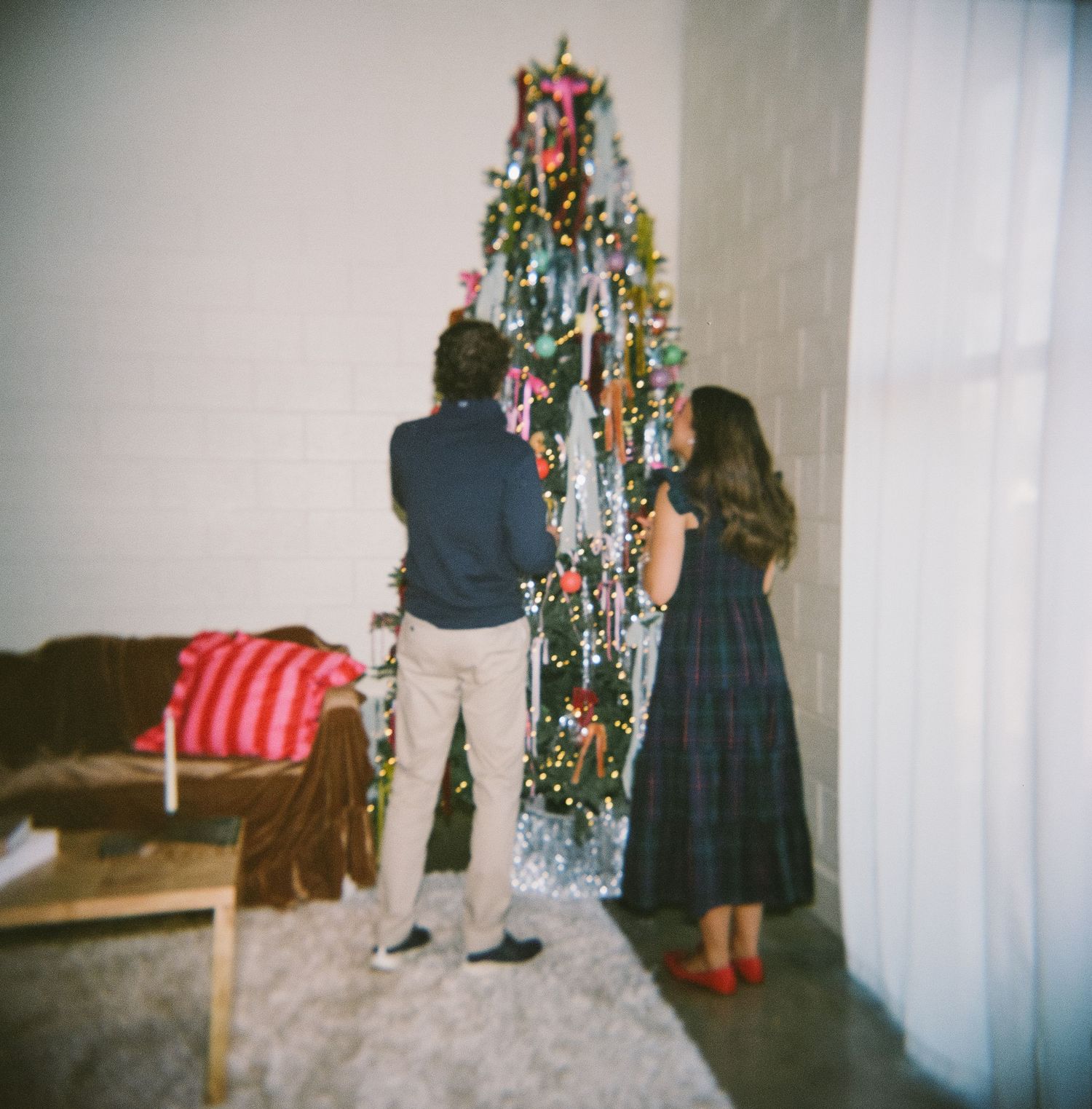 A couple stands facing a decorated Christmas tree with twinkling lights and ornaments.