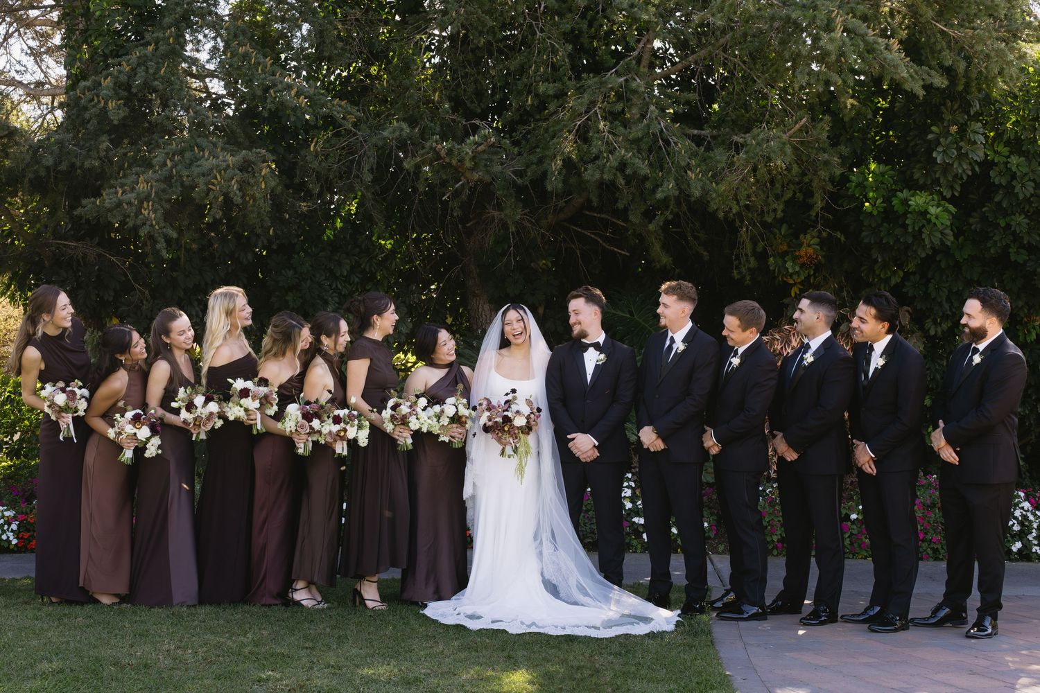 Wedding party lined up outdoors with bridesmaids in brown and groomsmen in black suits.