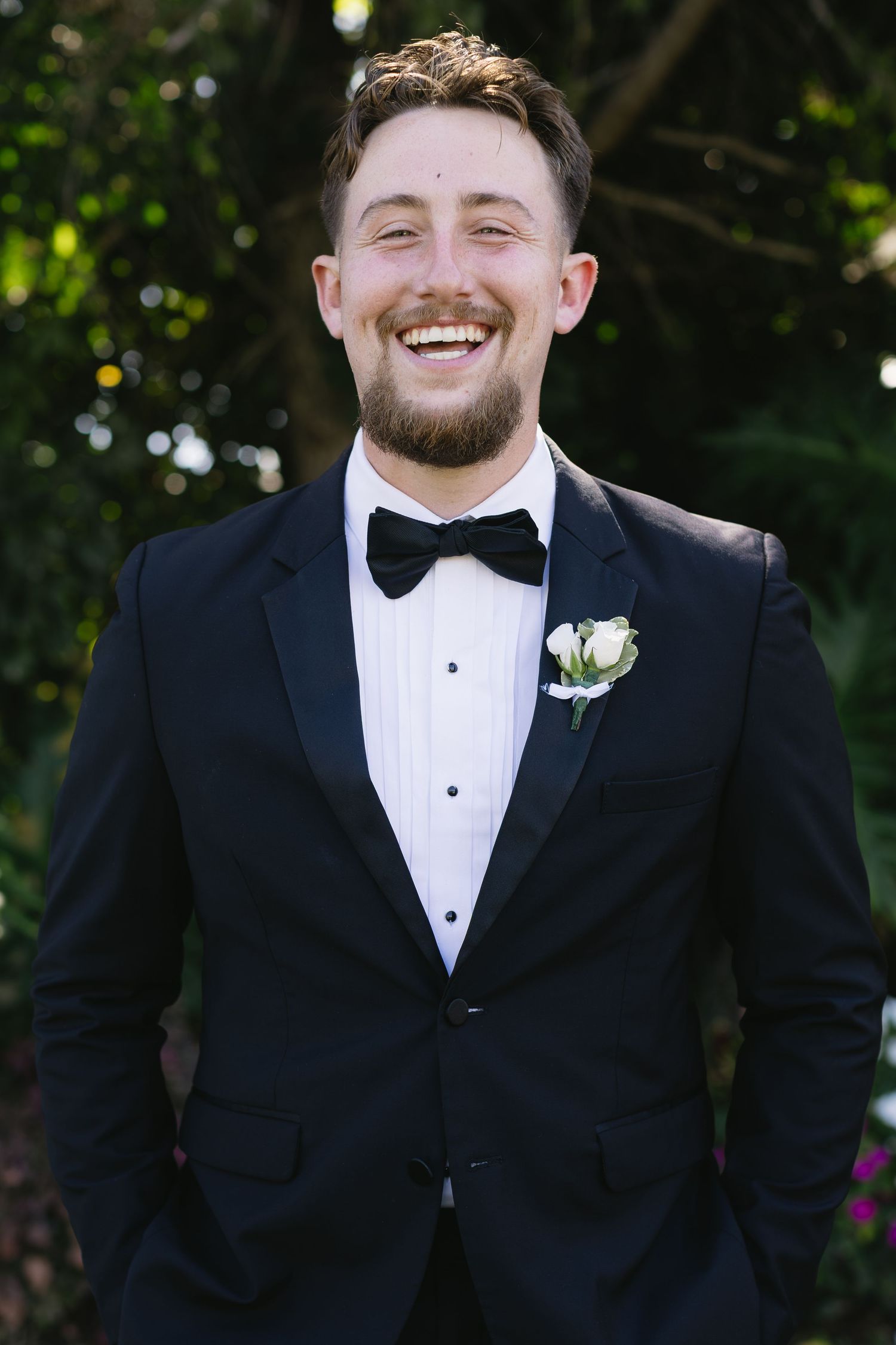 A groom in a classic black tuxedo and bowtie smiles joyfully during wedding portraits.