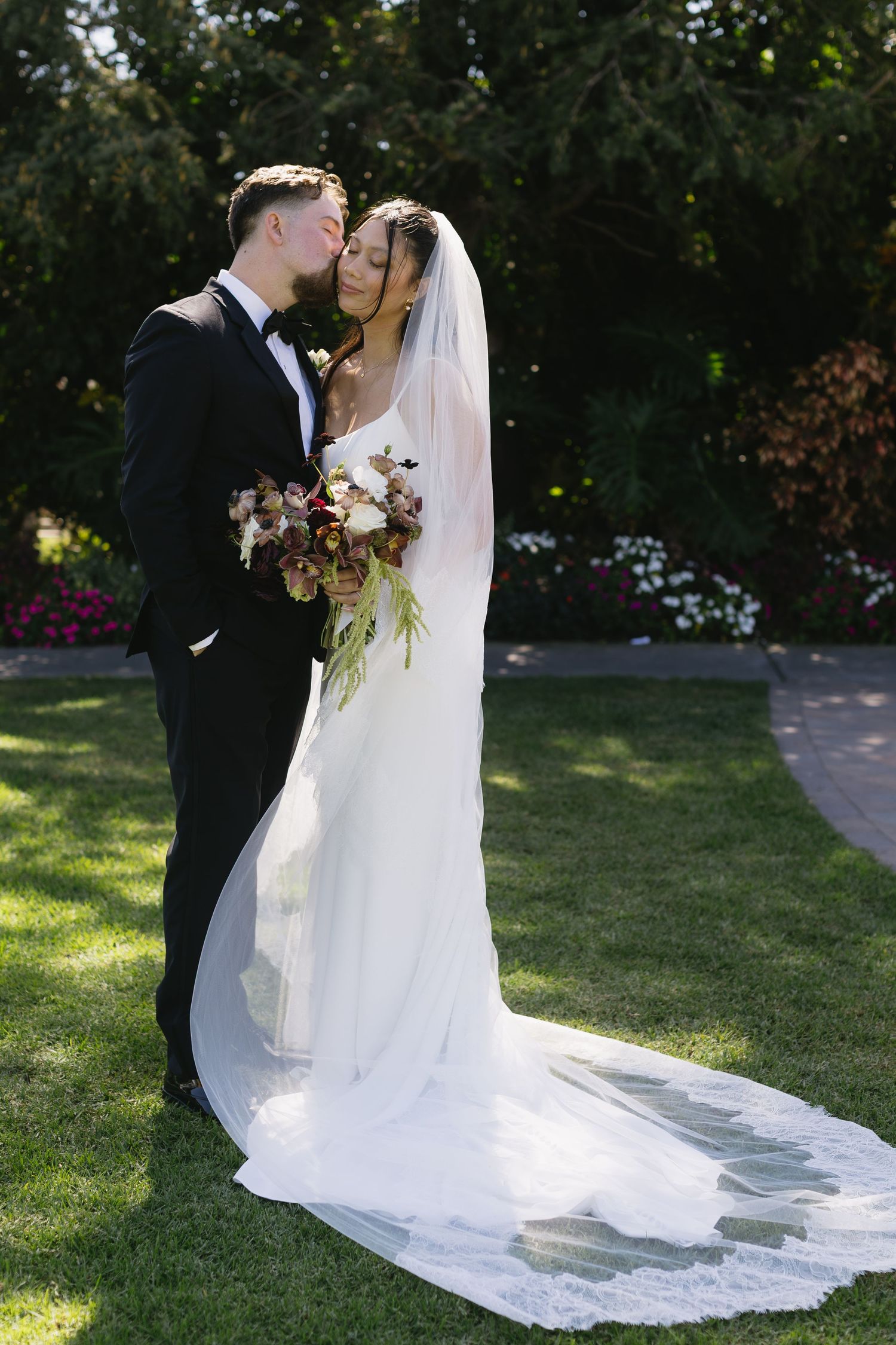 Romantic wedding couple embraces in a sunlit garden setting with flowing white veil and formal attire.