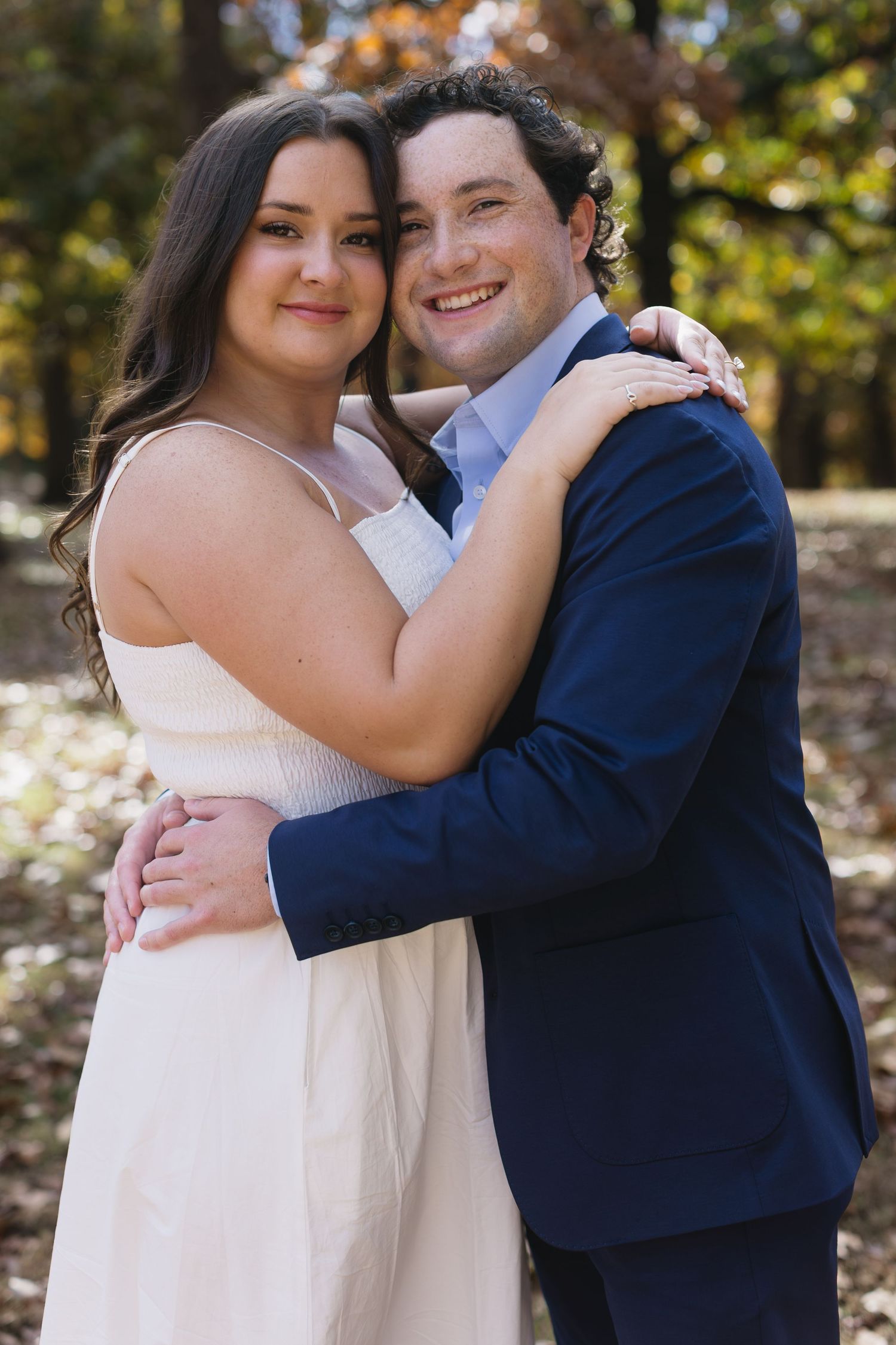 A couple in formal attire embraces outdoors during an autumn photoshoot with colorful foliage in the background.