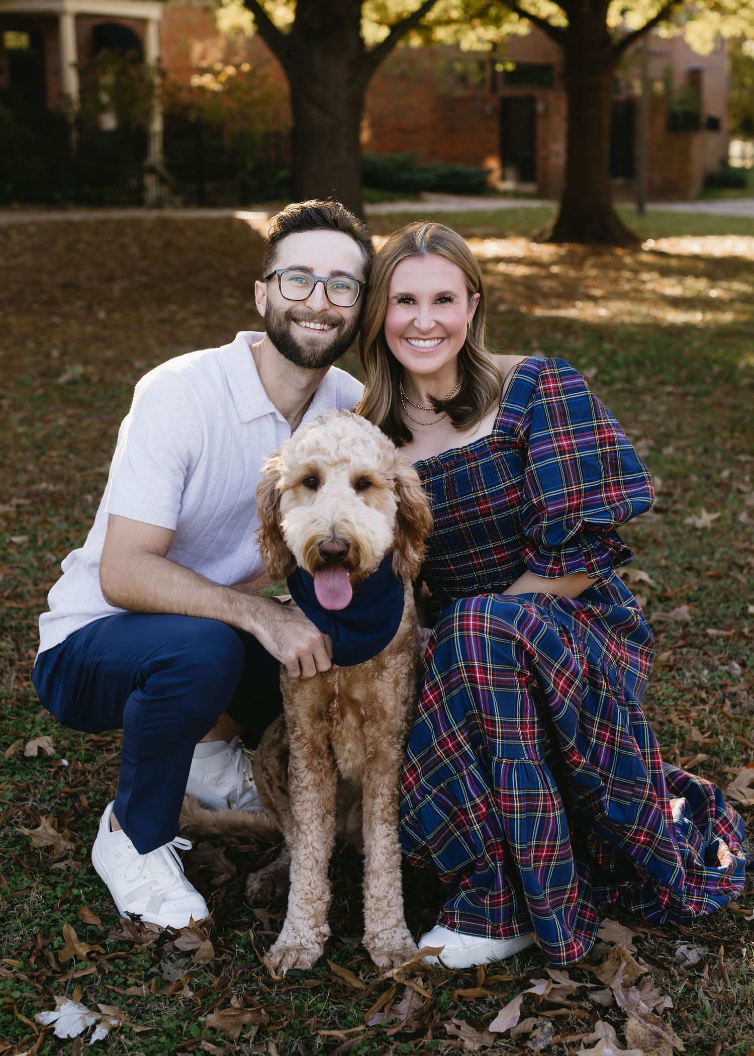 A couple poses with their goldendoodle dog wearing a navy bandana on an autumn day.