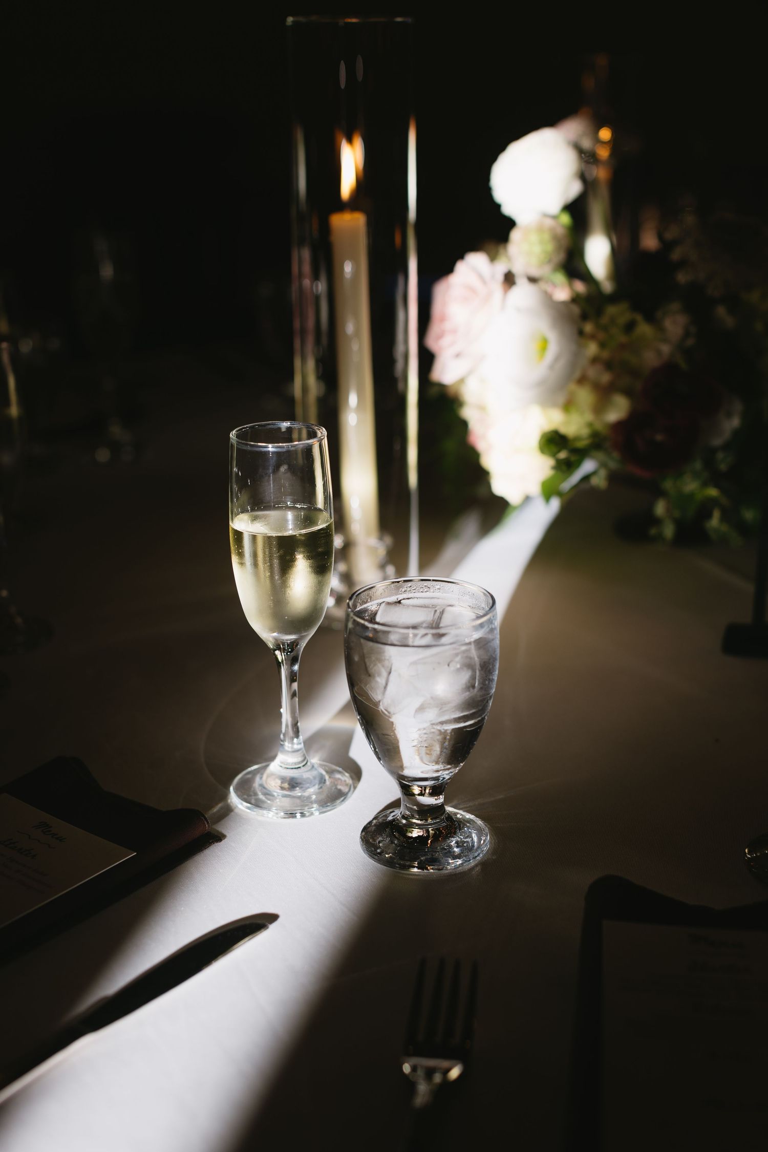 Elegant champagne glasses sit on a dark table with dramatic lighting and floral decorations.