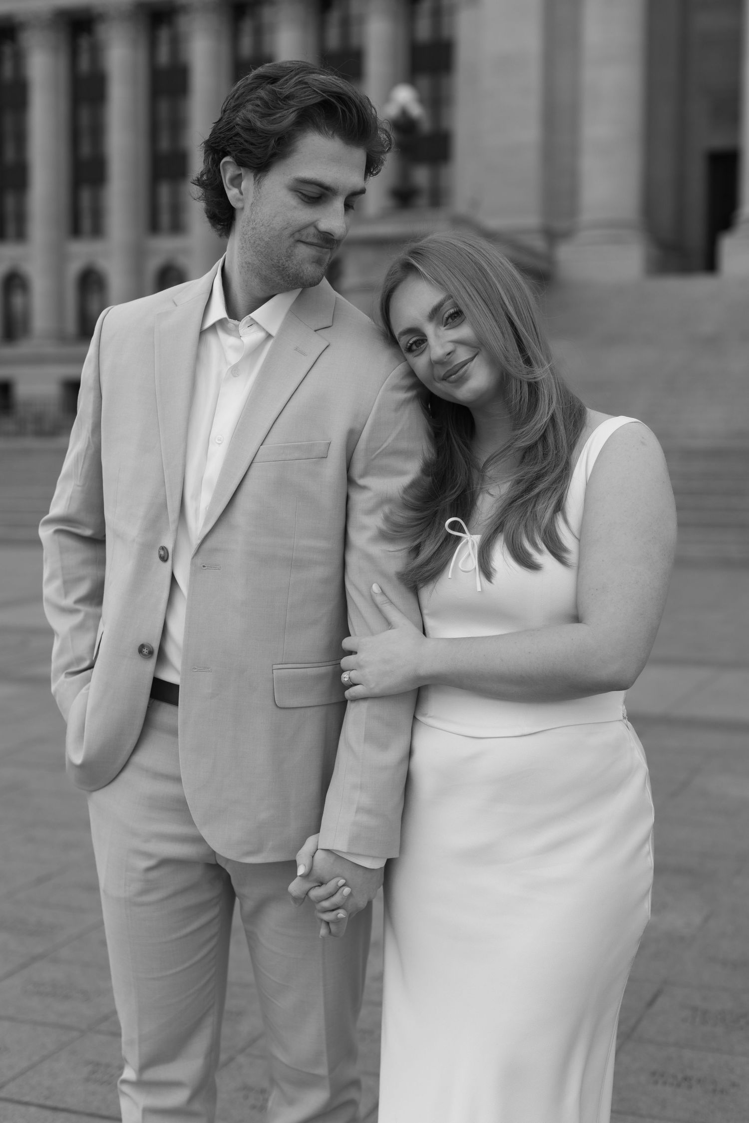 A couple in matching beige and white formal attire embrace while standing outside a building.