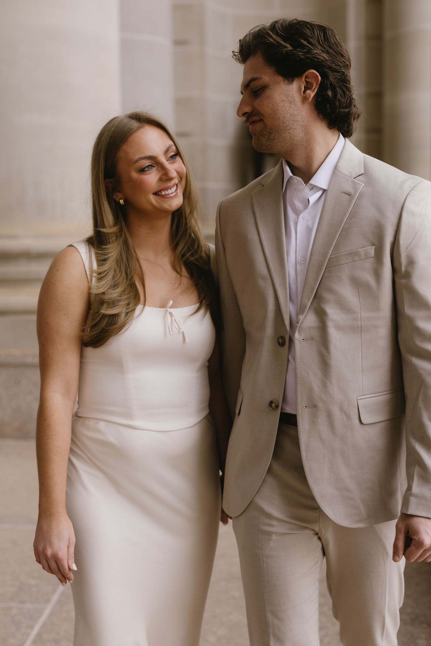 A romantic couple in coordinated beige and white formal wear posing against a classical building backdrop.