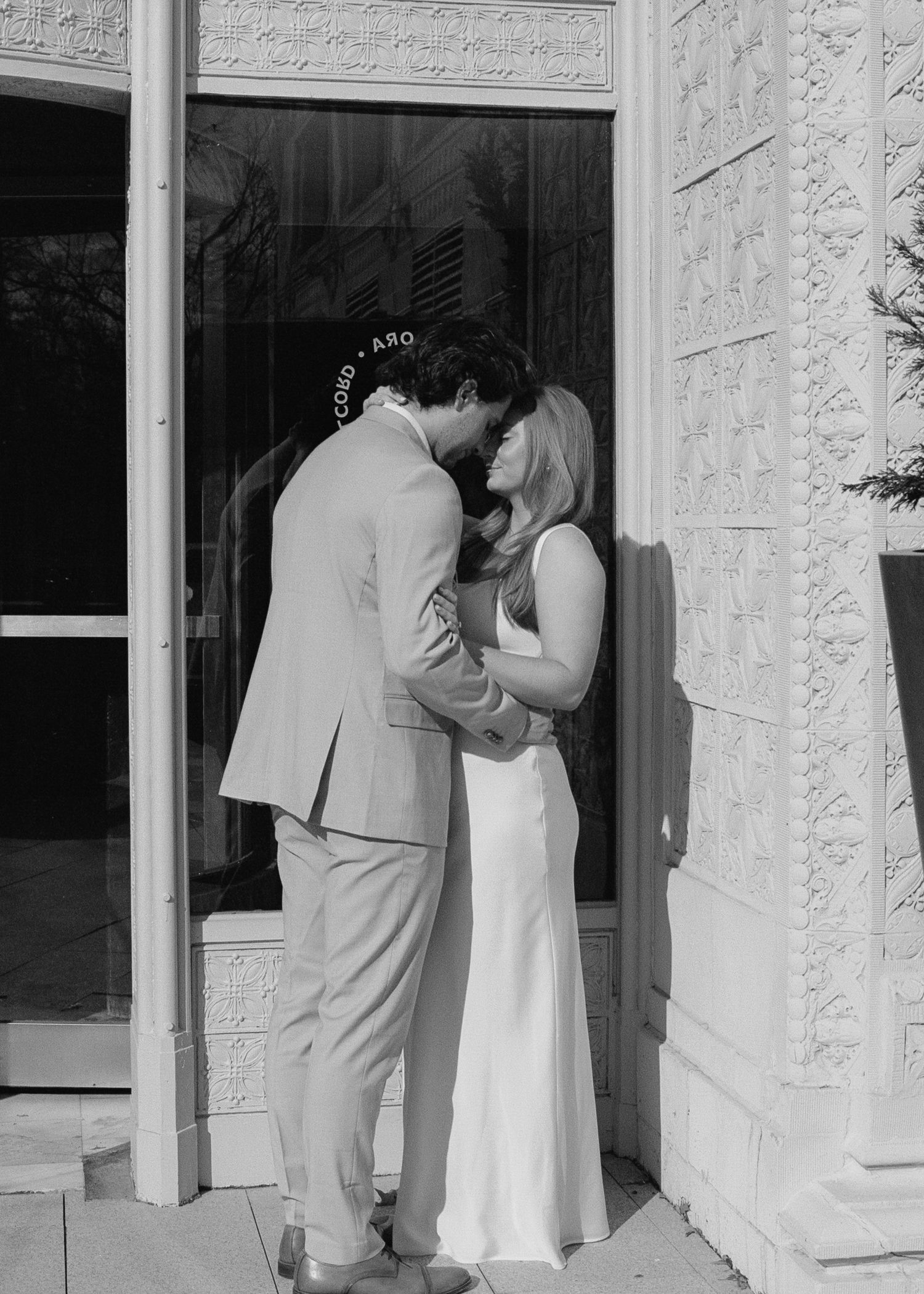 A tender moment between a couple in wedding attire against a white stone wall.