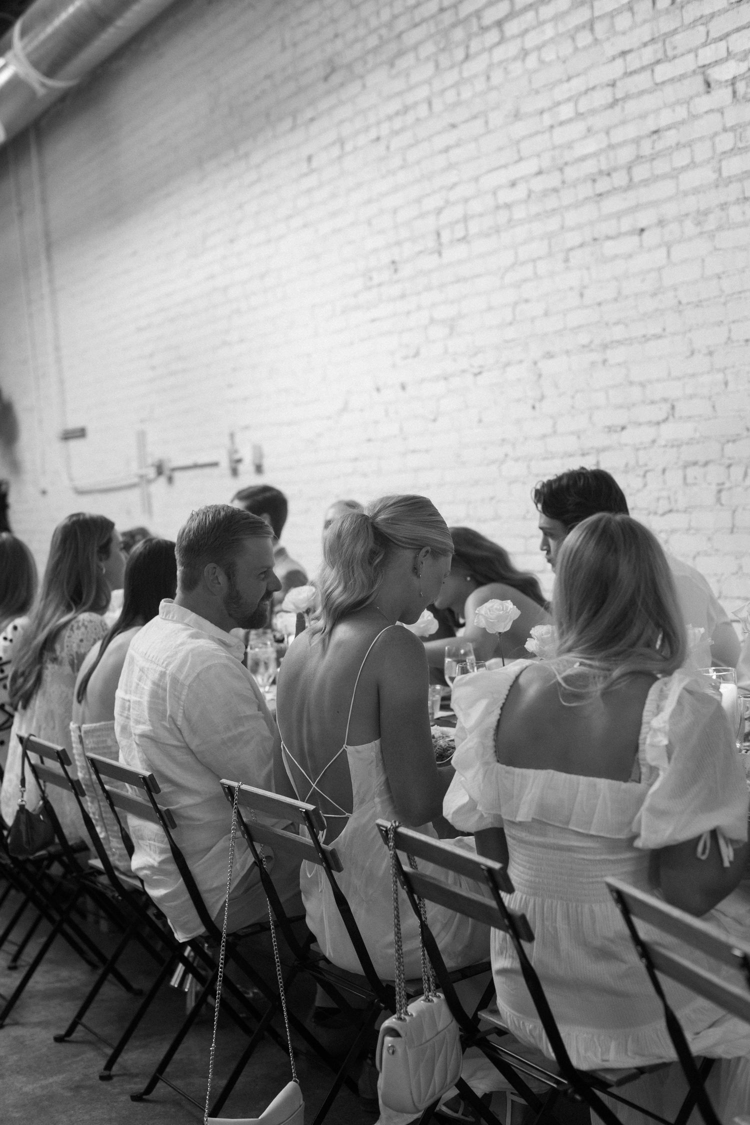 Black and white photo of guests seated at long tables in a white brick loft style venue.