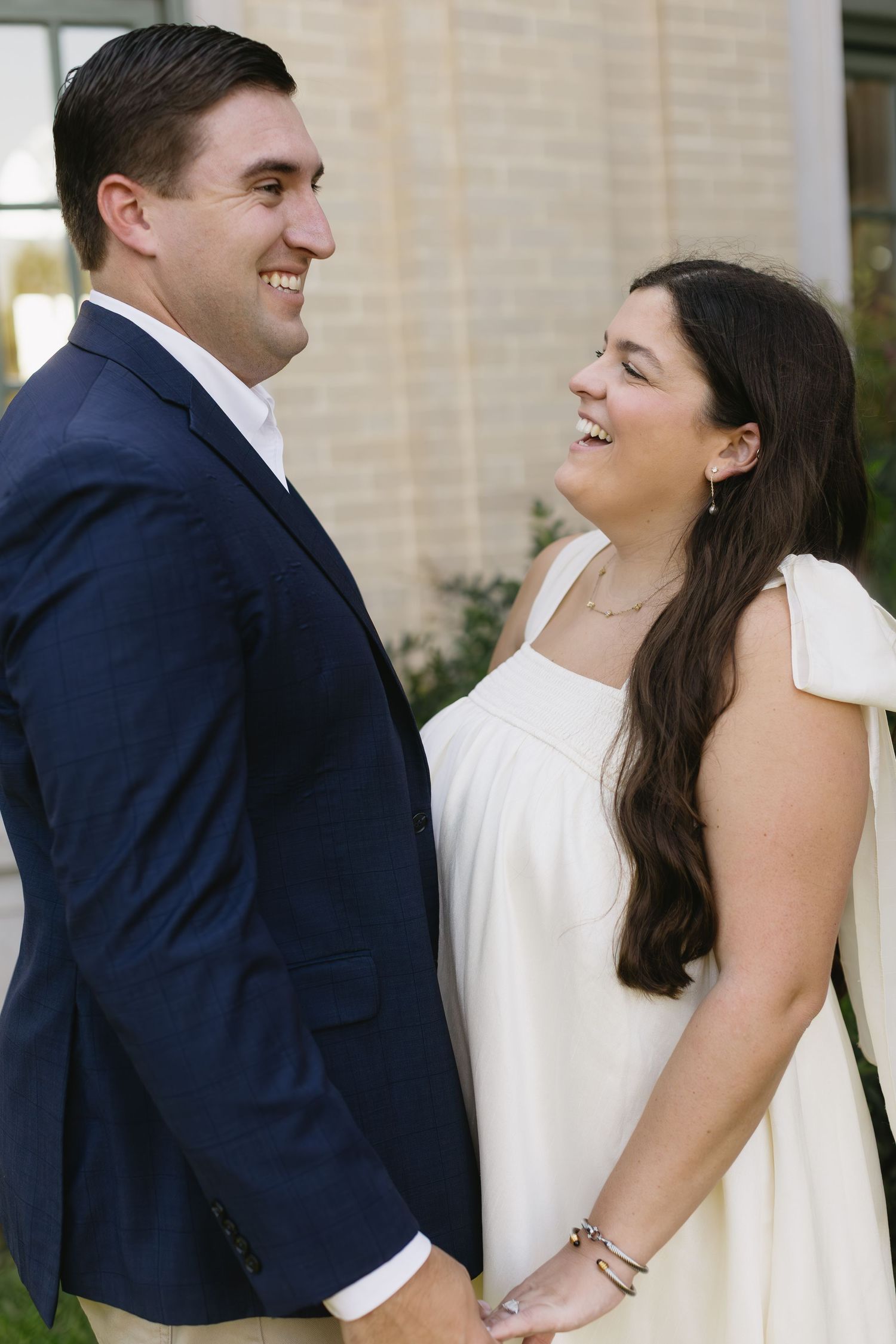 A couple shares intimate laughing moments in formal attire against a light building exterior.
