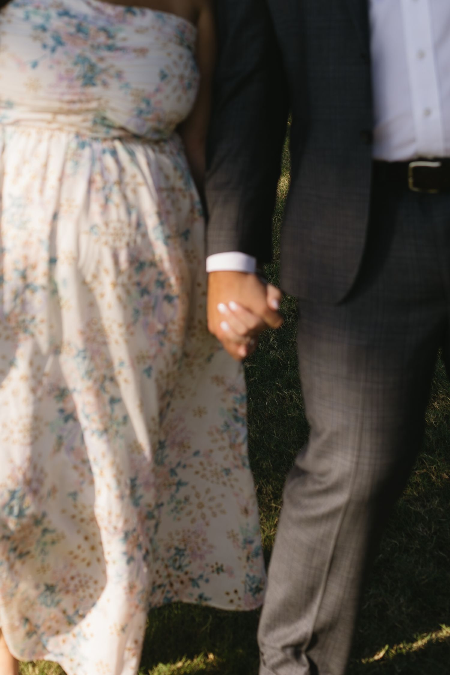 A couple in wedding attire stands together outdoors, with focus on a floral patterned dress and grey suit pants.