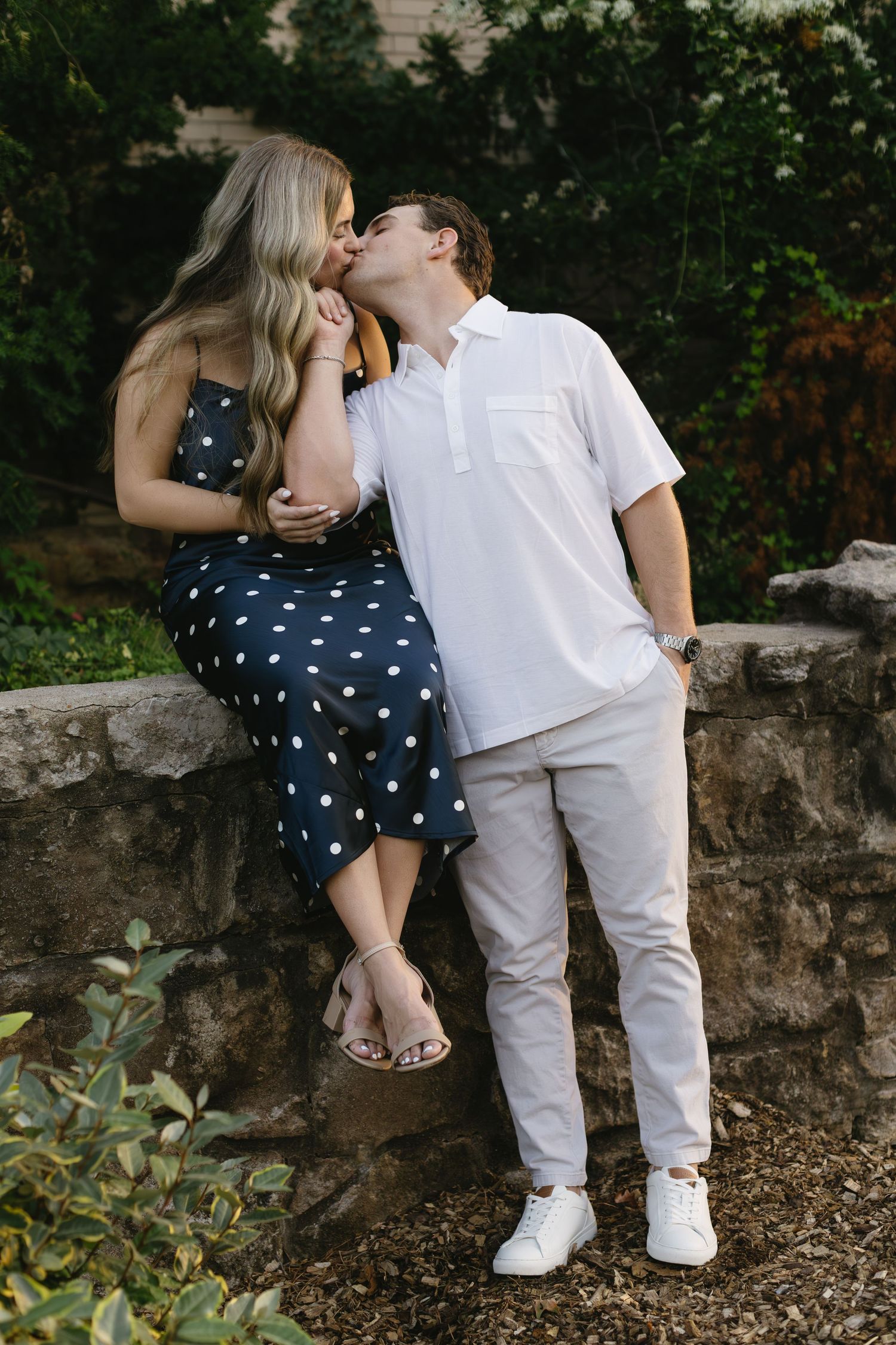 A couple shares an intimate moment outdoors, with her perched on a stone wall in a dotted dress.