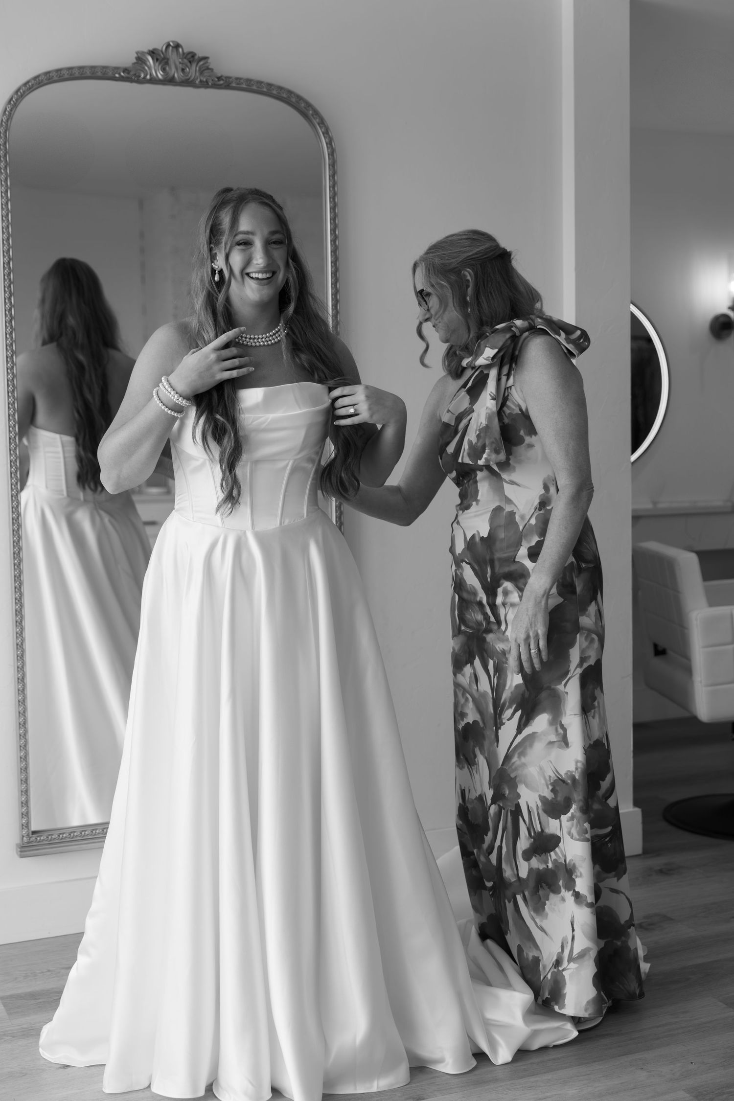 A candid moment showing a bride getting ready in a classic white wedding gown with someone in a floral dress helping.
