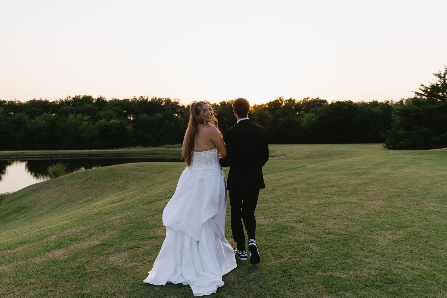 Romantic couple in wedding attire walking hand in hand across scenic outdoor venue at sunset.