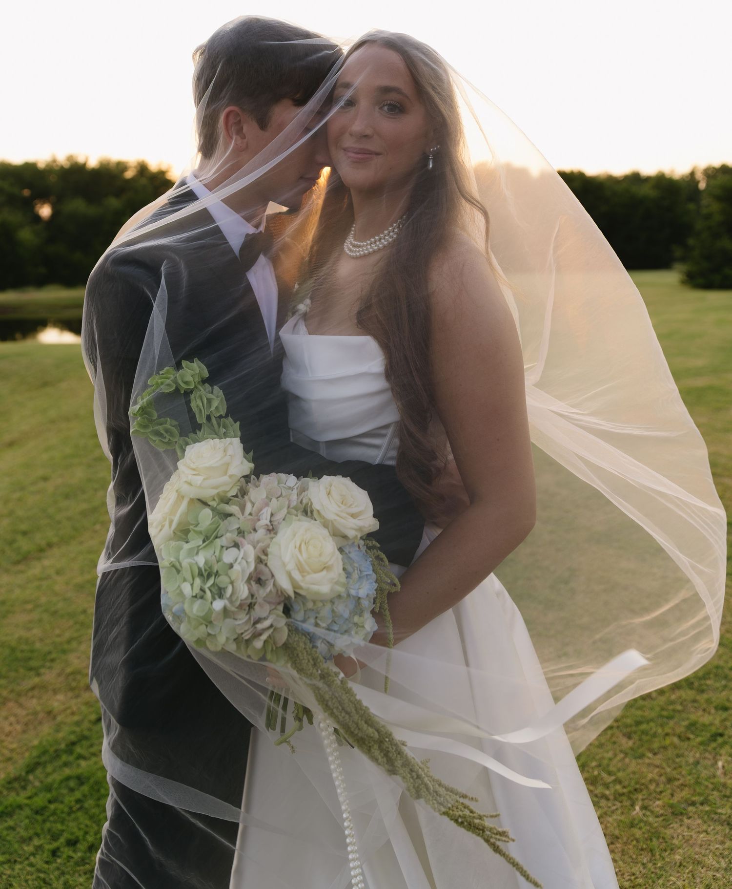 Wedding couple embraces outdoors at sunset with bride holding a white floral bouquet.