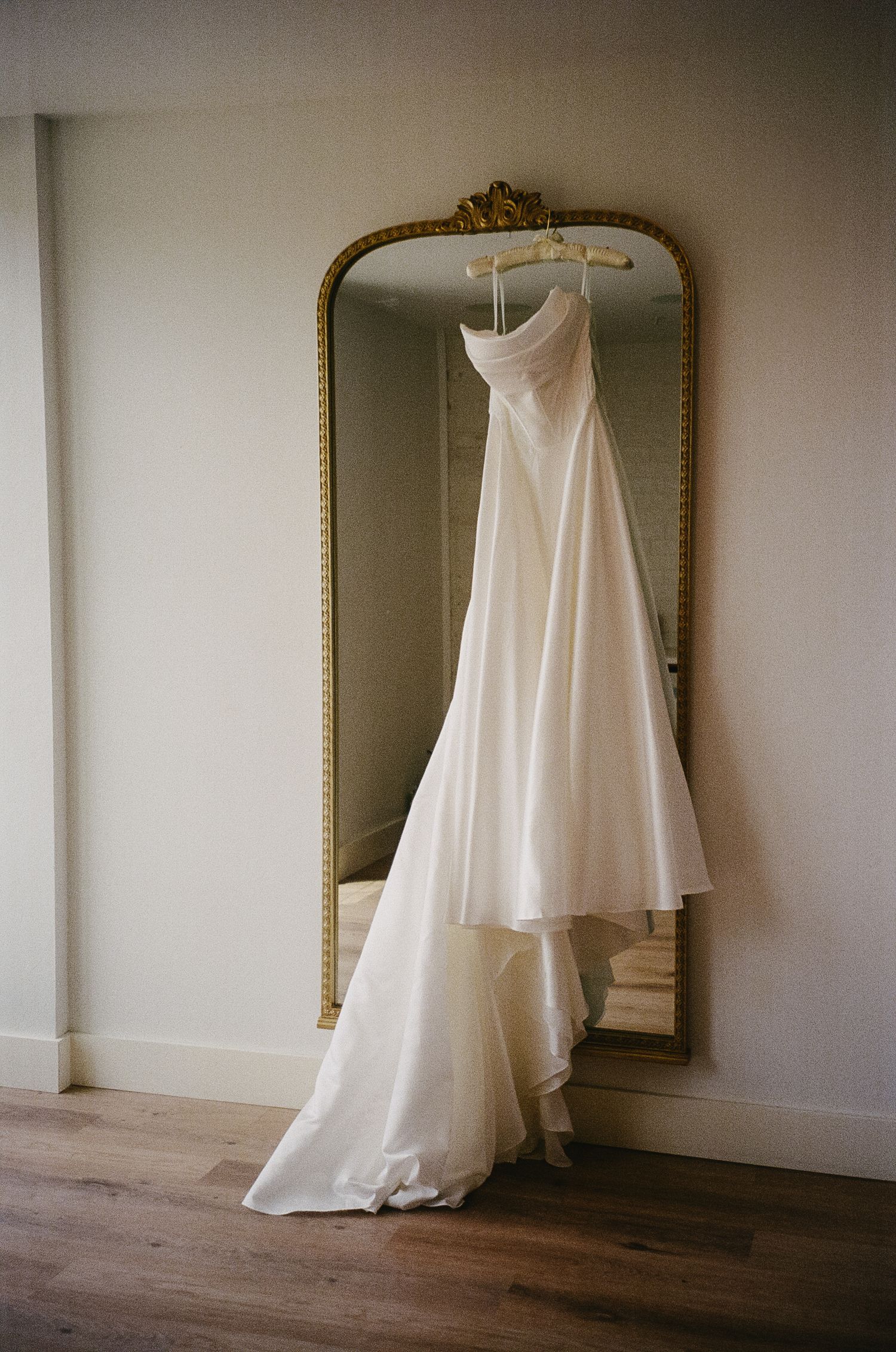 Elegant white wedding gown hanging against an ornate gold-framed mirror in a minimalist room.