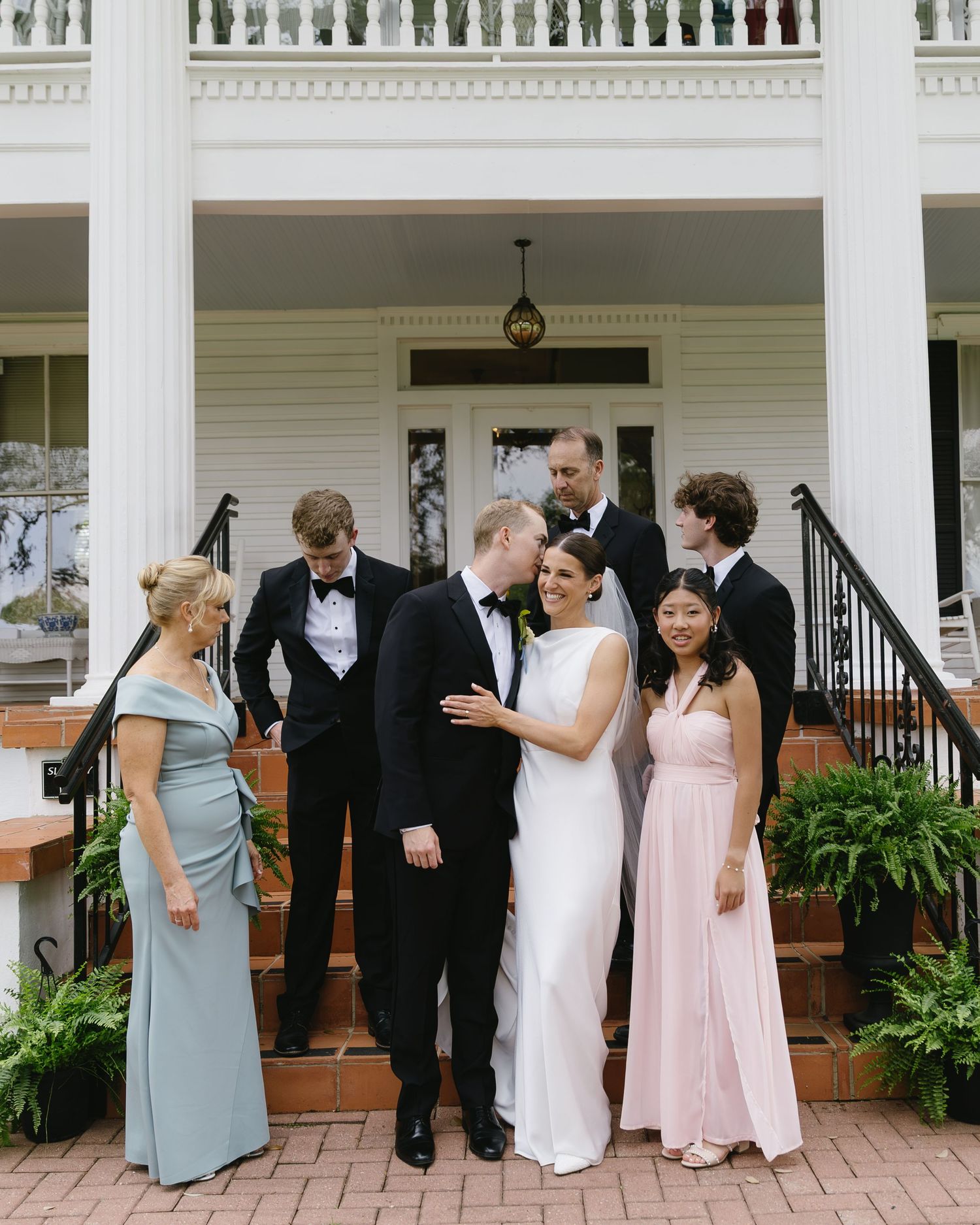 Wedding party gathered on white columned porch wearing formal attire.