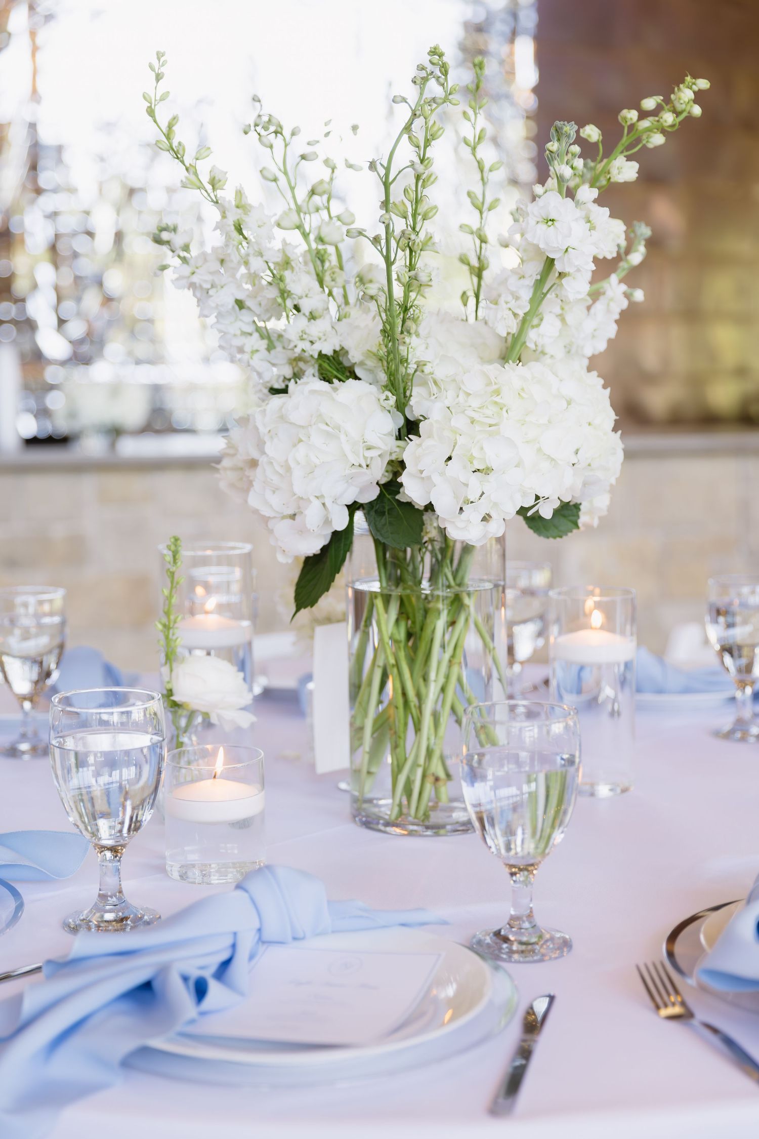 Elegant white floral centerpiece with delphinium and hydrangeas in a glass vase on a formal table setting.