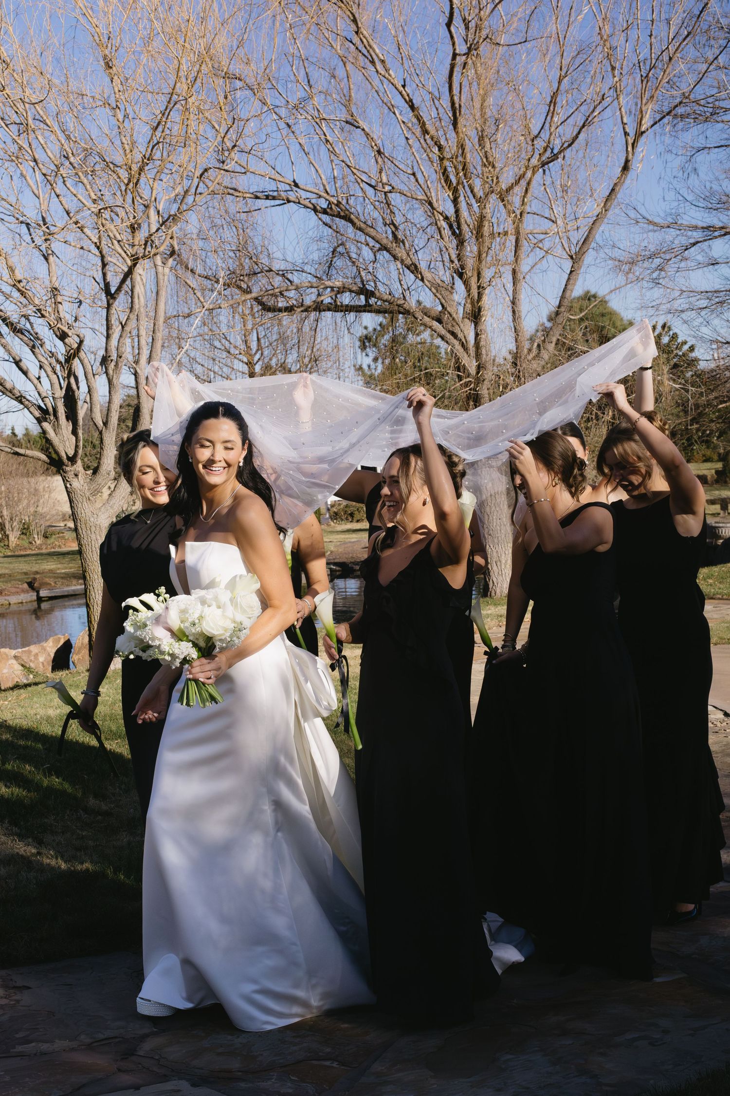 A bride in a white dress holds a bouquet while bridesmaids in black dresses play with her veil in an outdoor setting.
