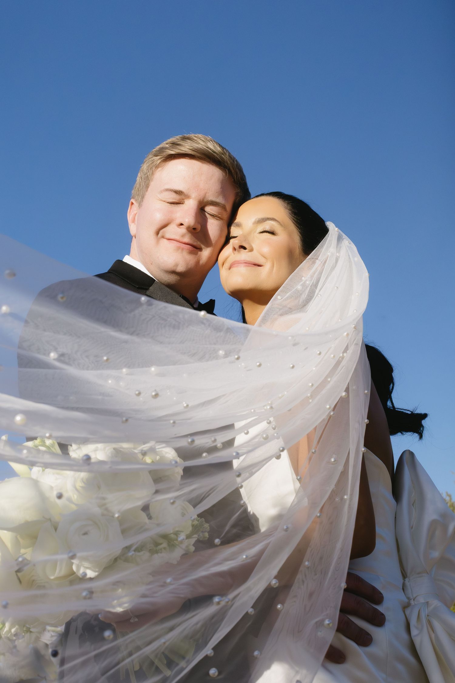 A romantic wedding portrait with flowing white veil against a bright blue sky background.