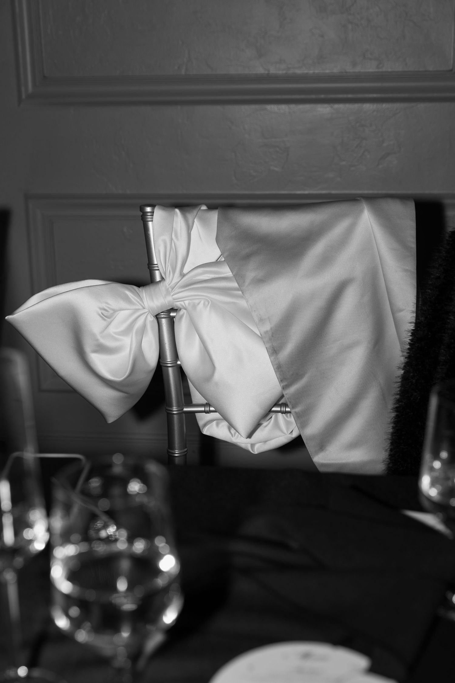 Black and white photo of white satin bow decoration with wine glass in foreground.