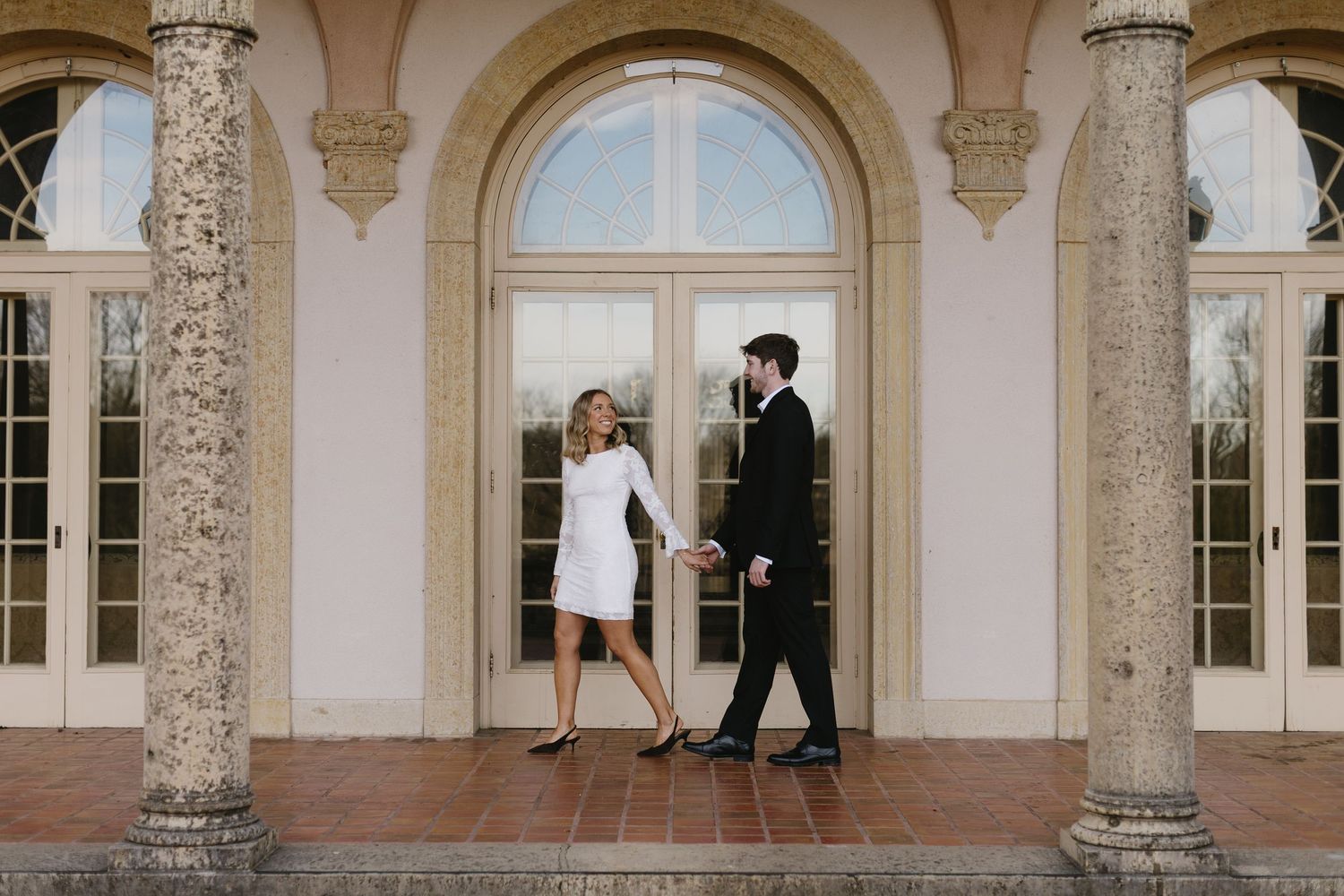 Elegant stone columns frame a walkway at a historic mansion with people visible through arched doorways.