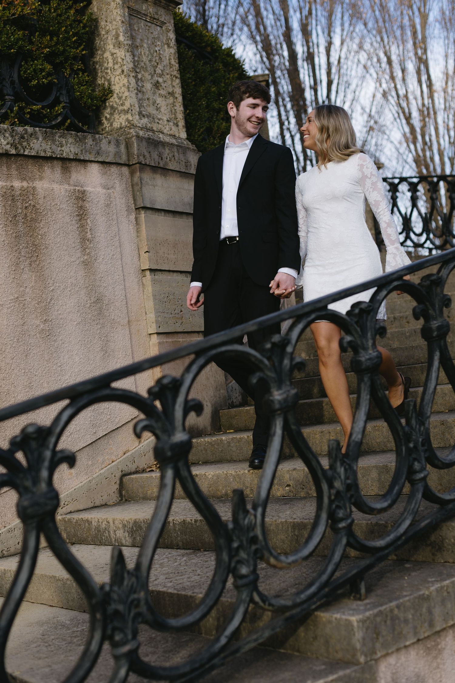 A couple stands near an ornate black iron railing with bare trees in the background.