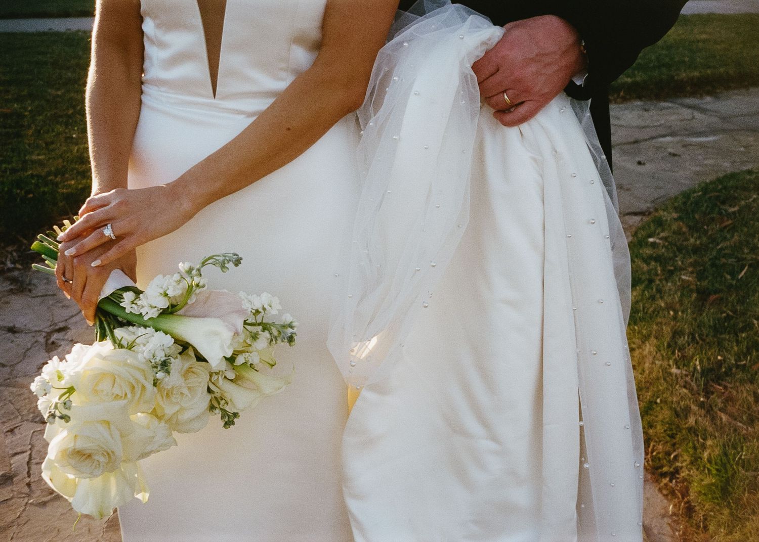Close-up details of bride's white wedding dress and floral bouquet outdoors.