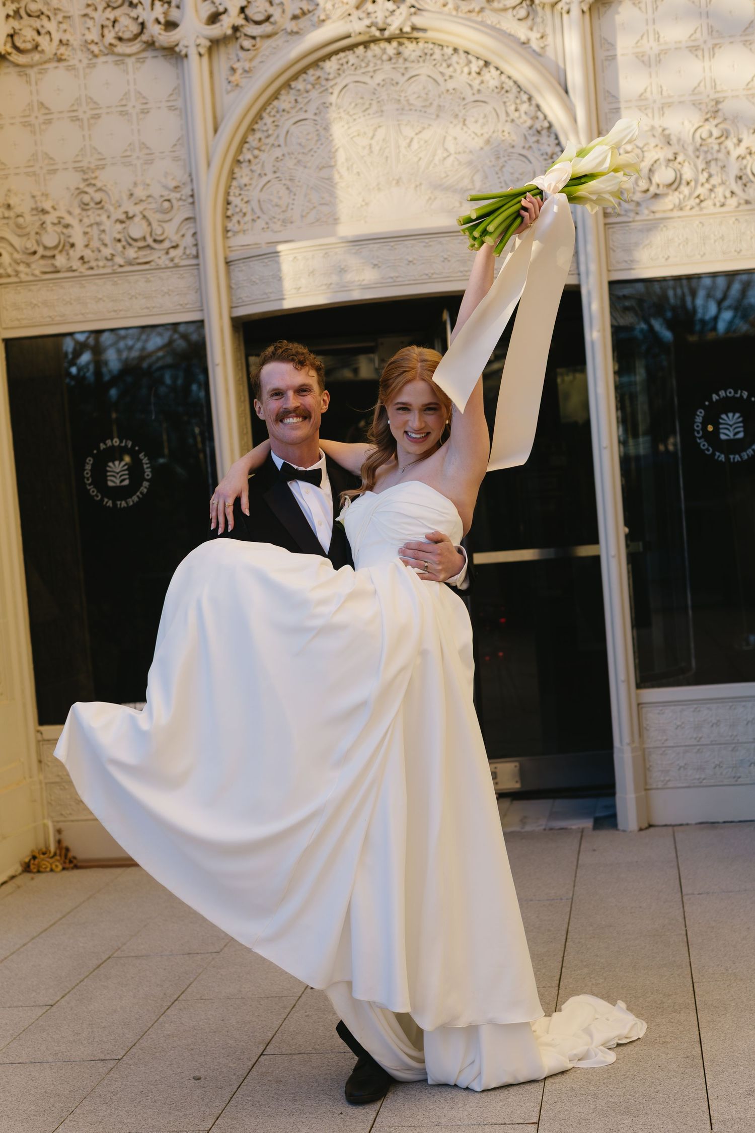 Couple in formal wedding attire posing under an ornate white gazebo.