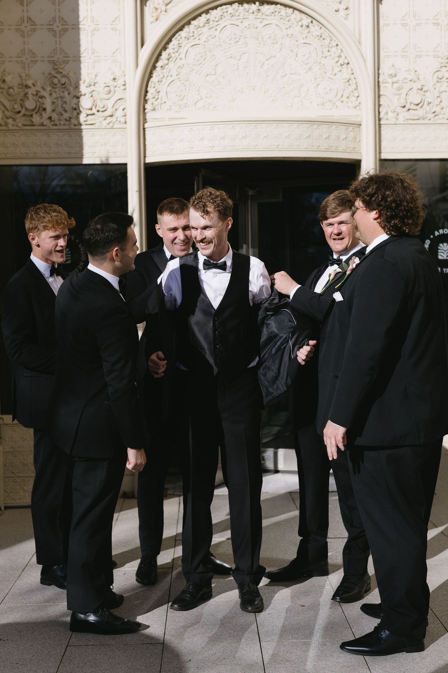 Groomsmen in black tuxedos line up against an ornate stone wall for wedding photos.