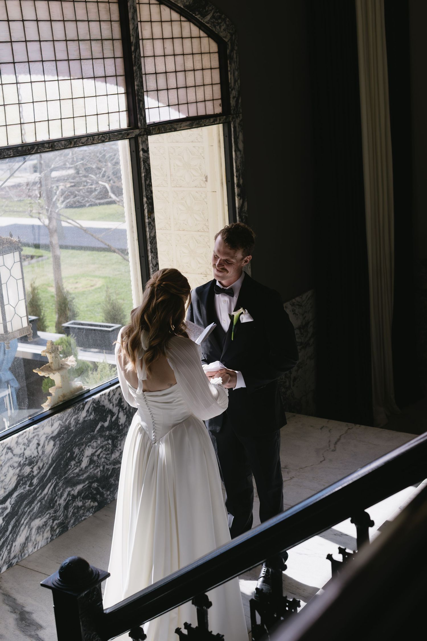 Bride in white dress and groom in tuxedo share a romantic moment by large windows.