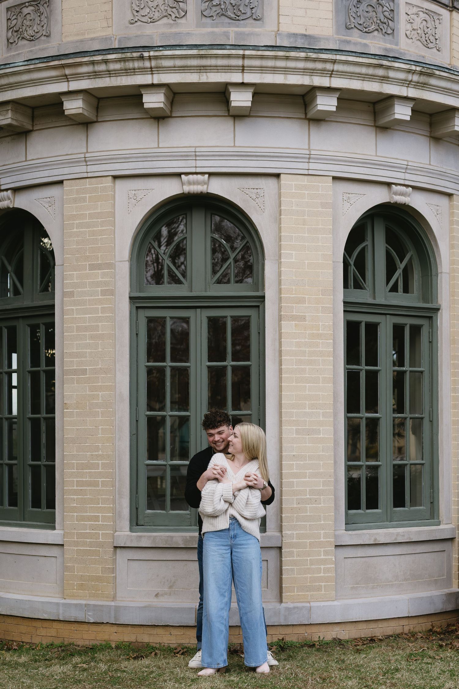 A couple embraces in front of an elegant stone building with arched windows.