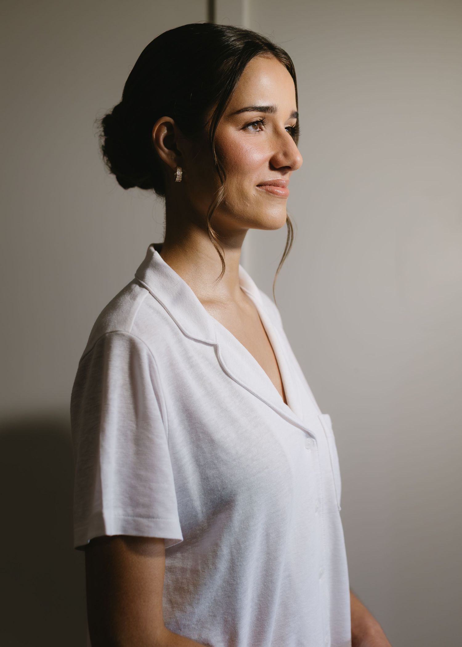 A woman in a casual white v-neck t-shirt poses against a neutral wall in a minimalist setting.