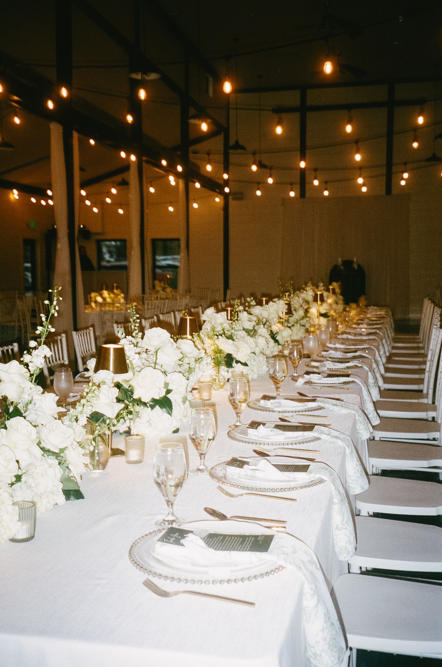 Elegant wedding reception tables set with white linens and floral centerpieces under string lights.