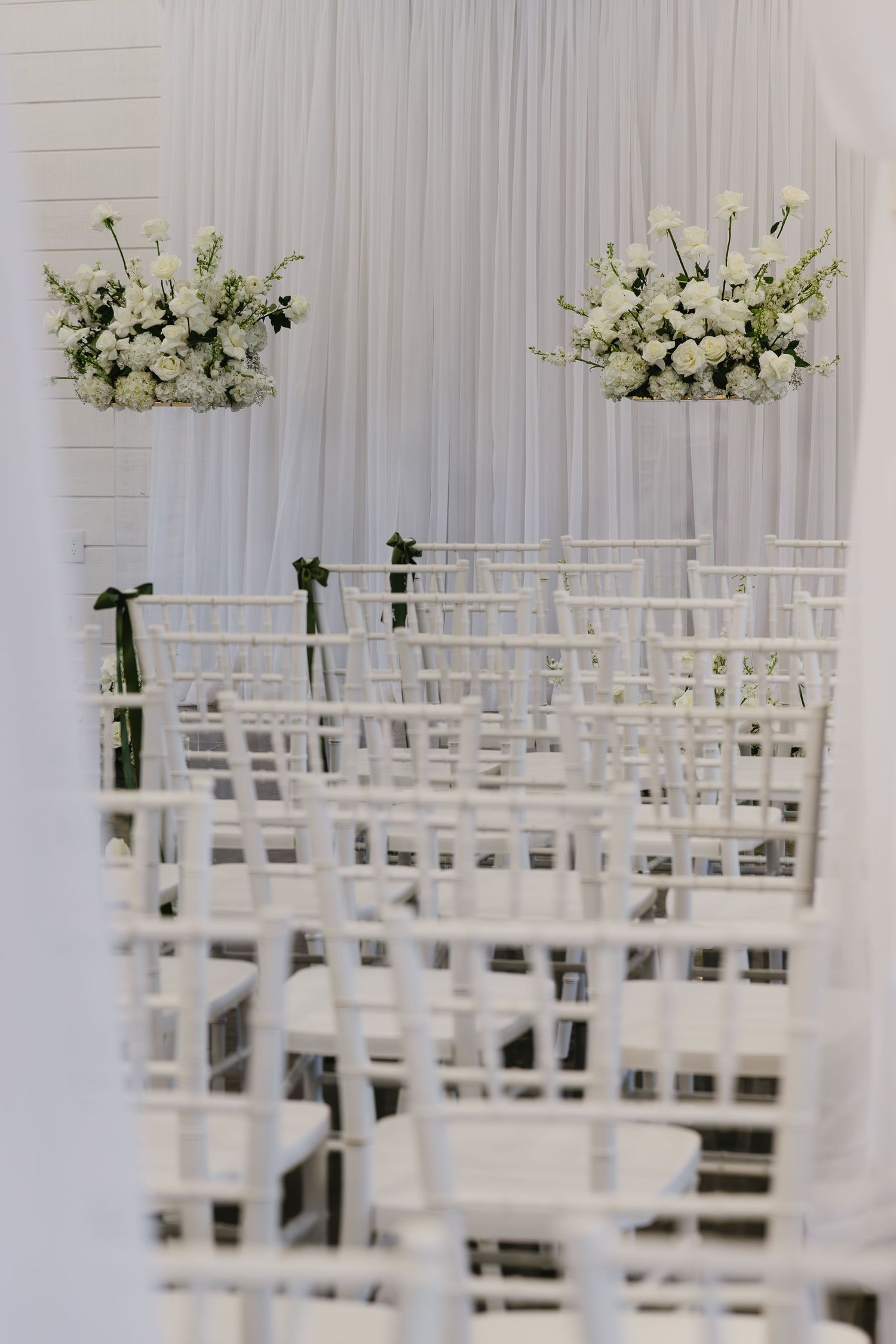 White chairs and floral arrangements set up for an elegant indoor wedding ceremony.