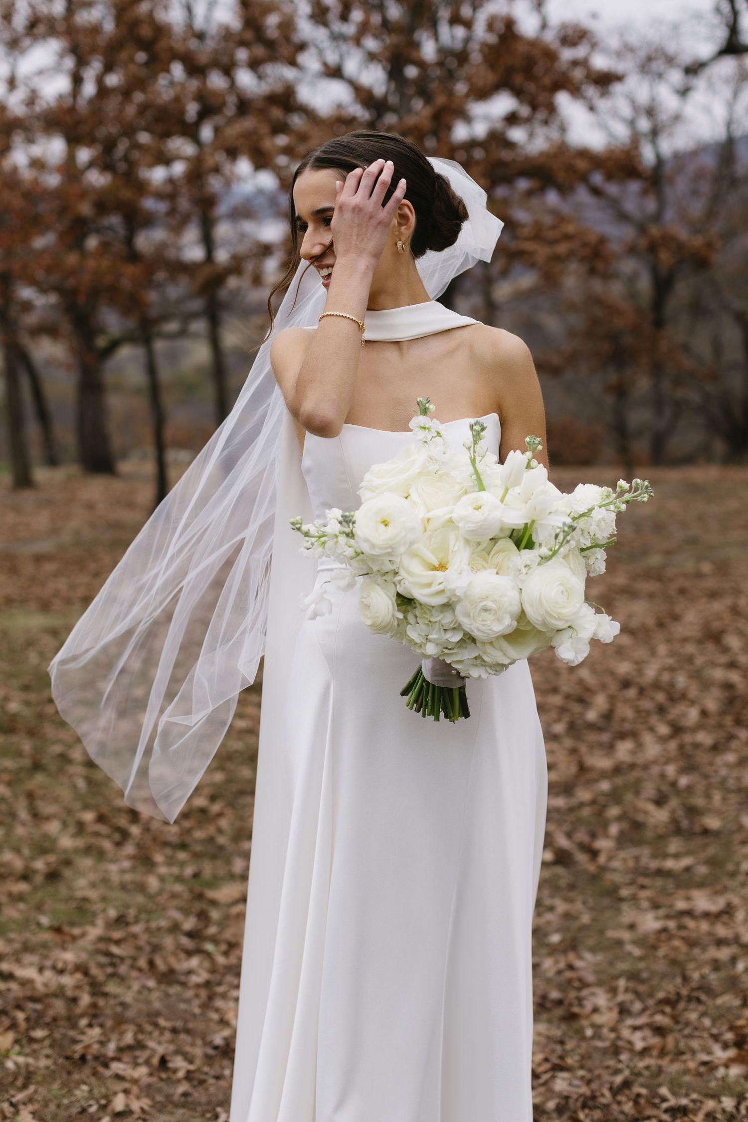 Bride in flowing white gown holds luxurious white bouquet against autumn landscape.