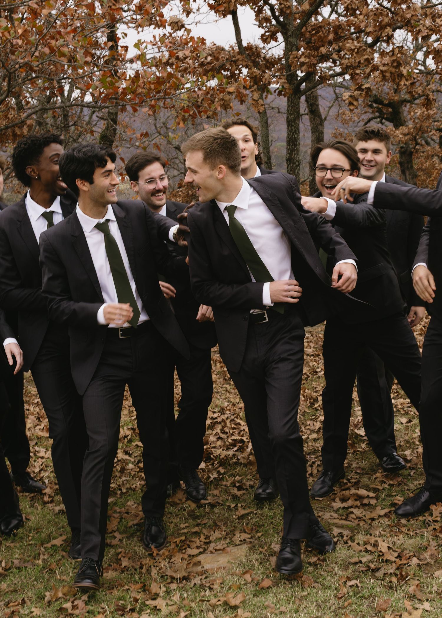 Wedding party groomsmen in black suits perform synchronized leaning poses outdoors.