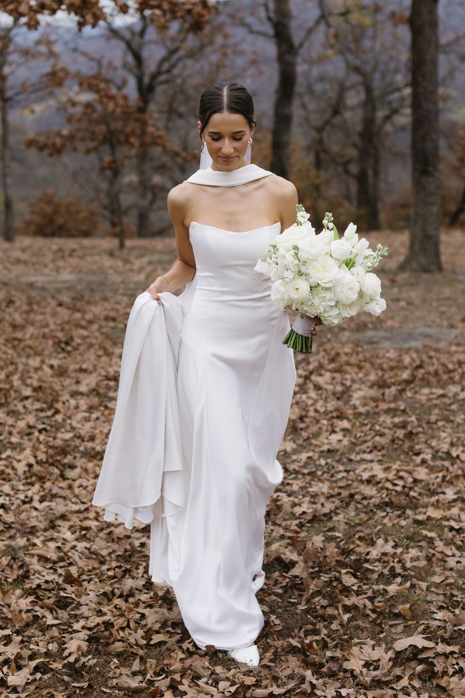 Elegant bridal portraits in a white gown holding white flowers against an autumn forest backdrop.