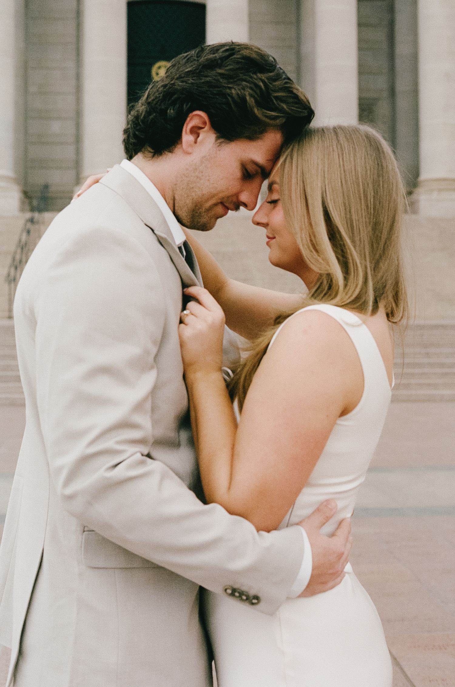 A romantic couple in light formal wear share an intimate moment in front of a grand building entrance.