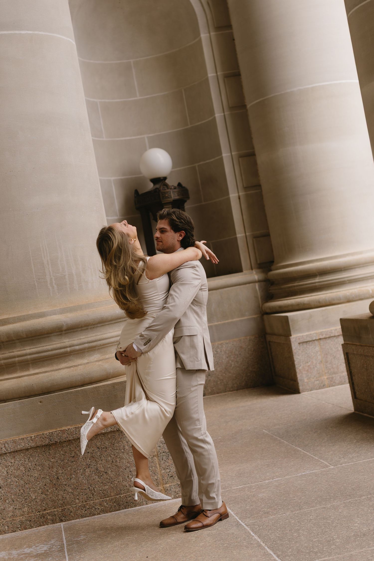 A couple in white attire shares a playful moment between elegant stone columns.