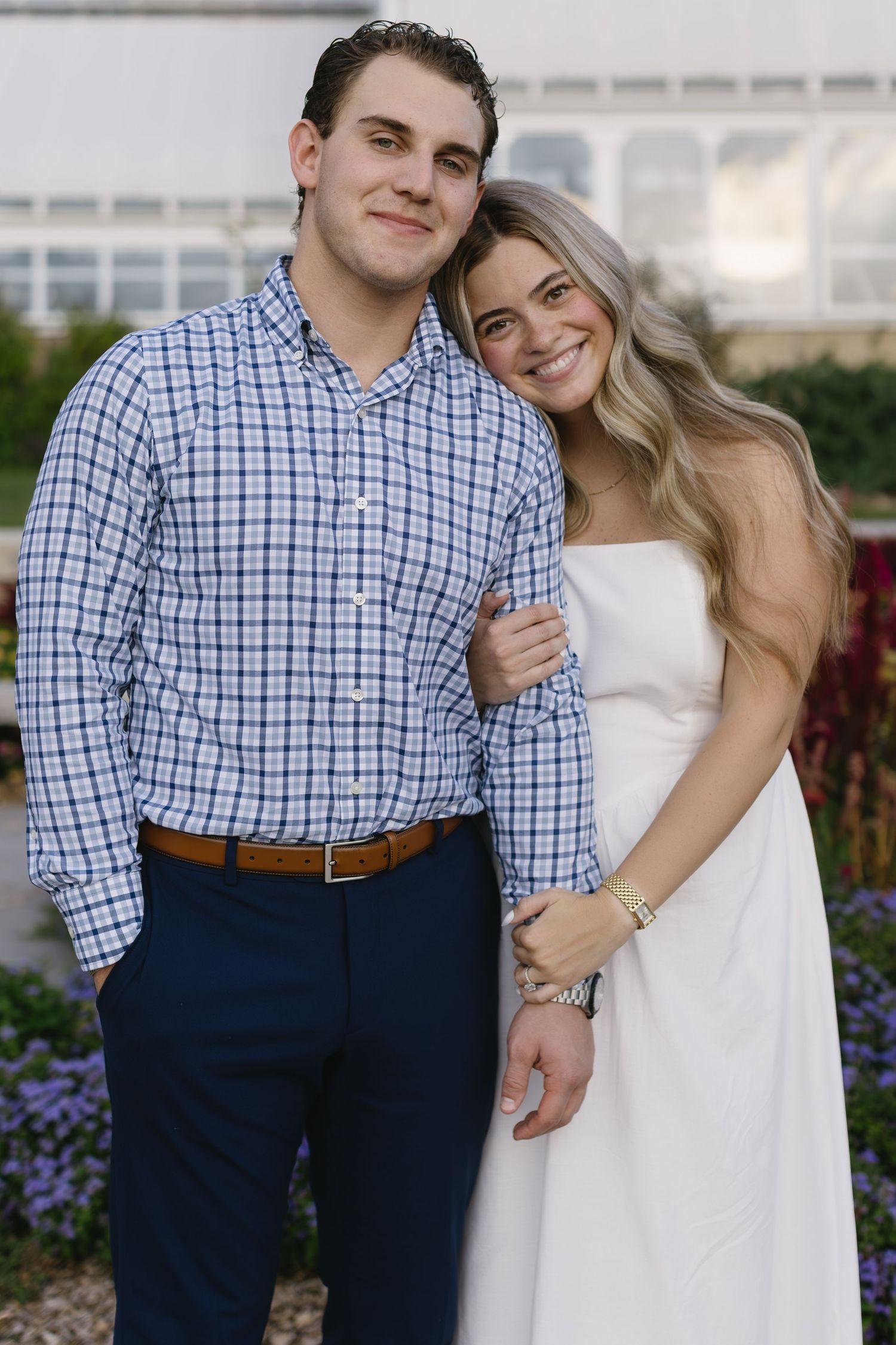 A couple smiles and embraces while dressed in formal attire with blue checkered shirt and white dress.