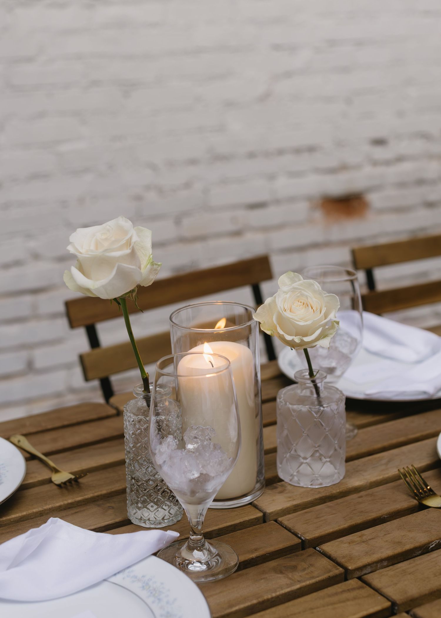 Elegant table setting with white roses in crystal vases and glowing candles on wooden slats.