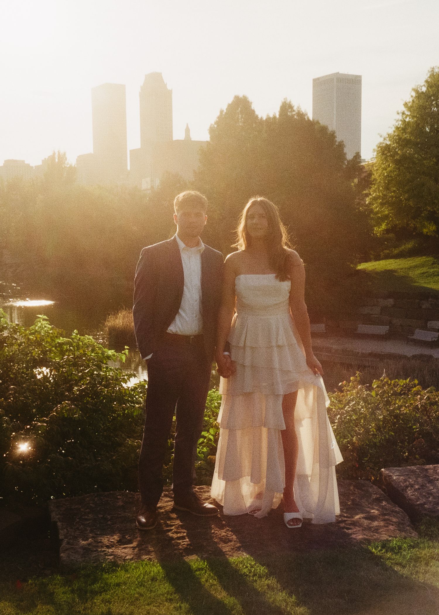A couple poses against a cityscape backdrop during sunset with golden light streaming through trees.