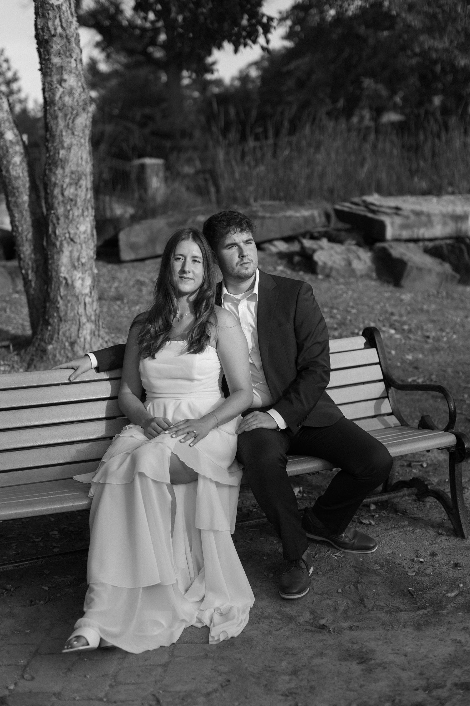 A black and white portrait of a couple in wedding attire sitting together on a park bench.