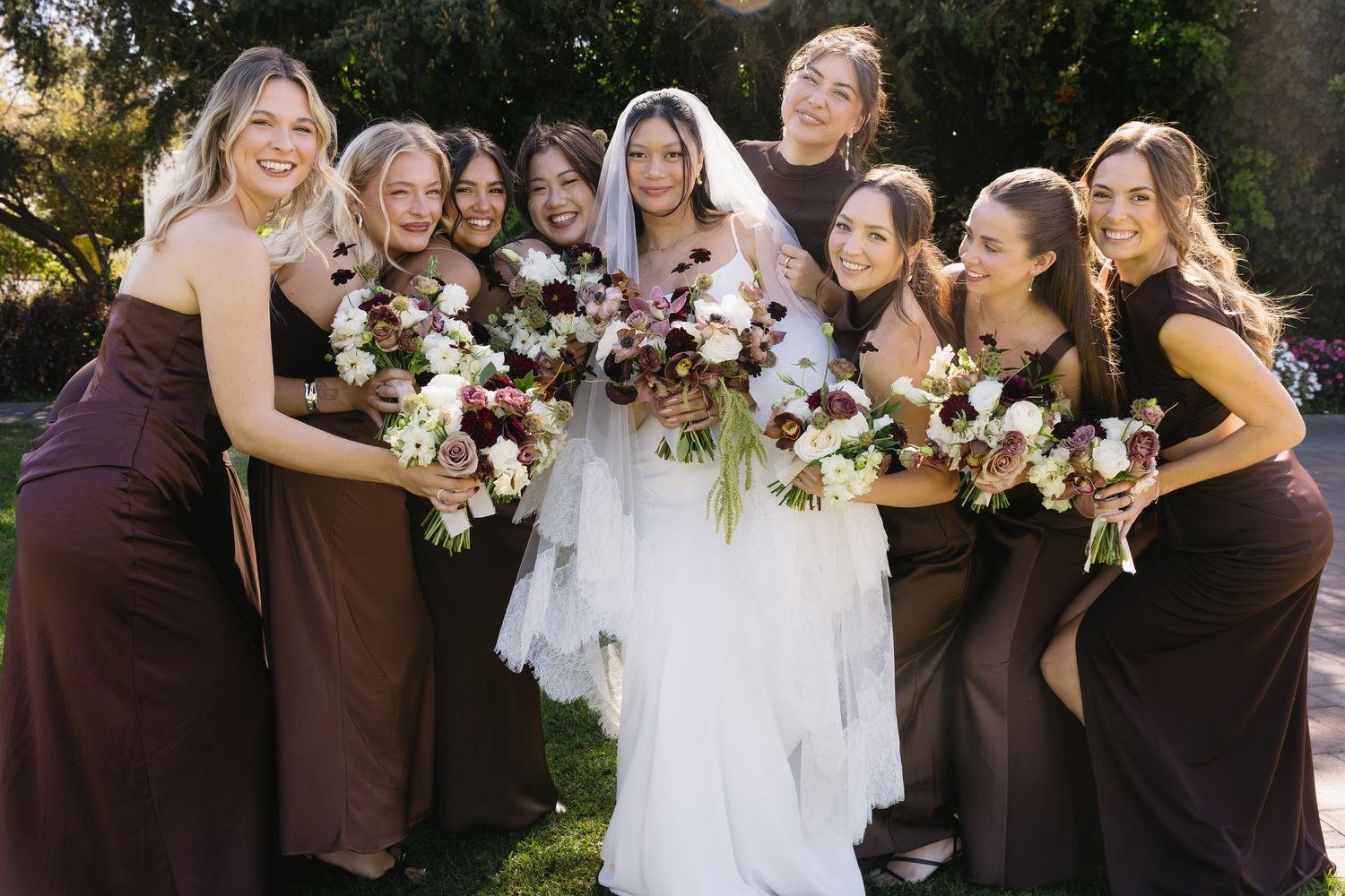 Wedding party in brown dresses and white gowns posing together outdoors.