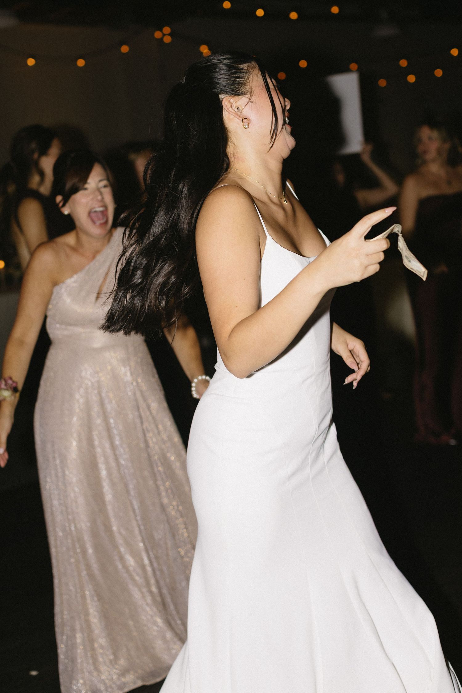 Wedding guests dancing and celebrating at a dimly lit reception with glowing lights in the background.